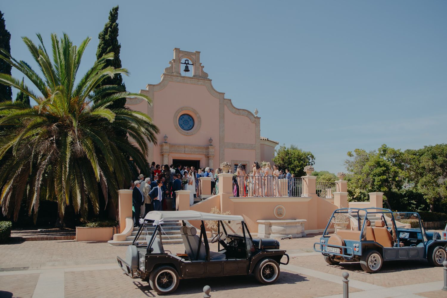 Pink Mediterranean-style church exterior with palm trees and vintage cars parked outside.