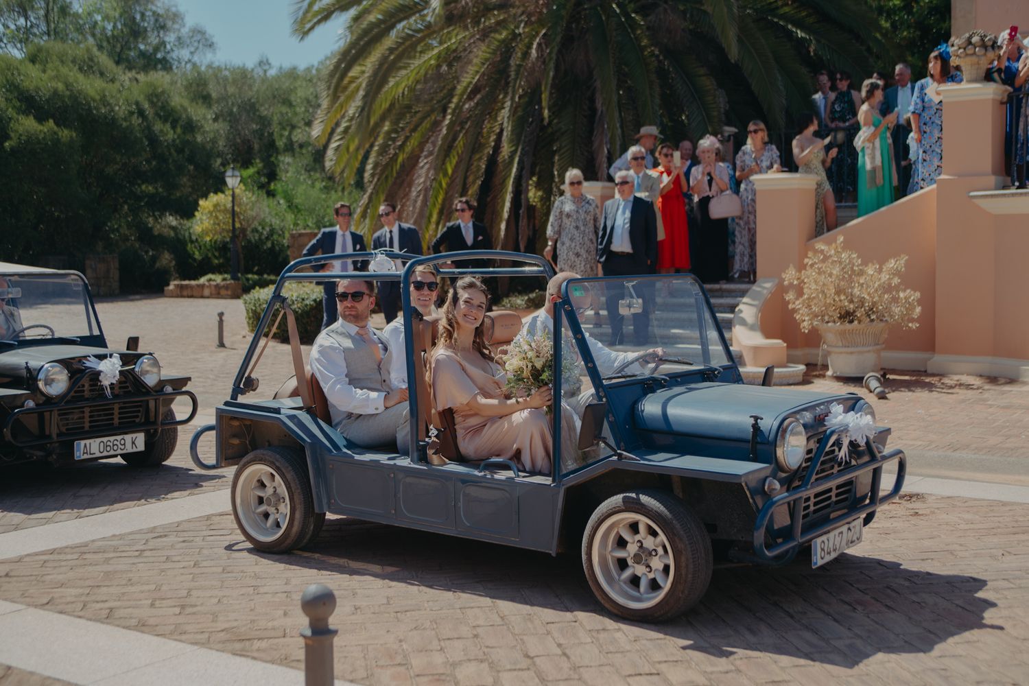 Wedding party rides in vintage blue convertible cars outside Mediterranean-style building.
