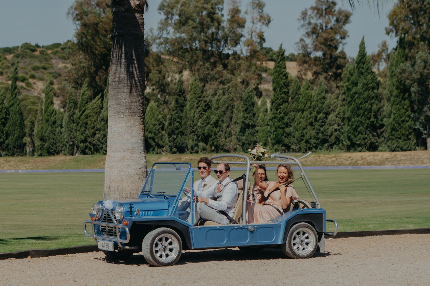 A vintage blue mini moke car parked on grass with wedding guests inside.