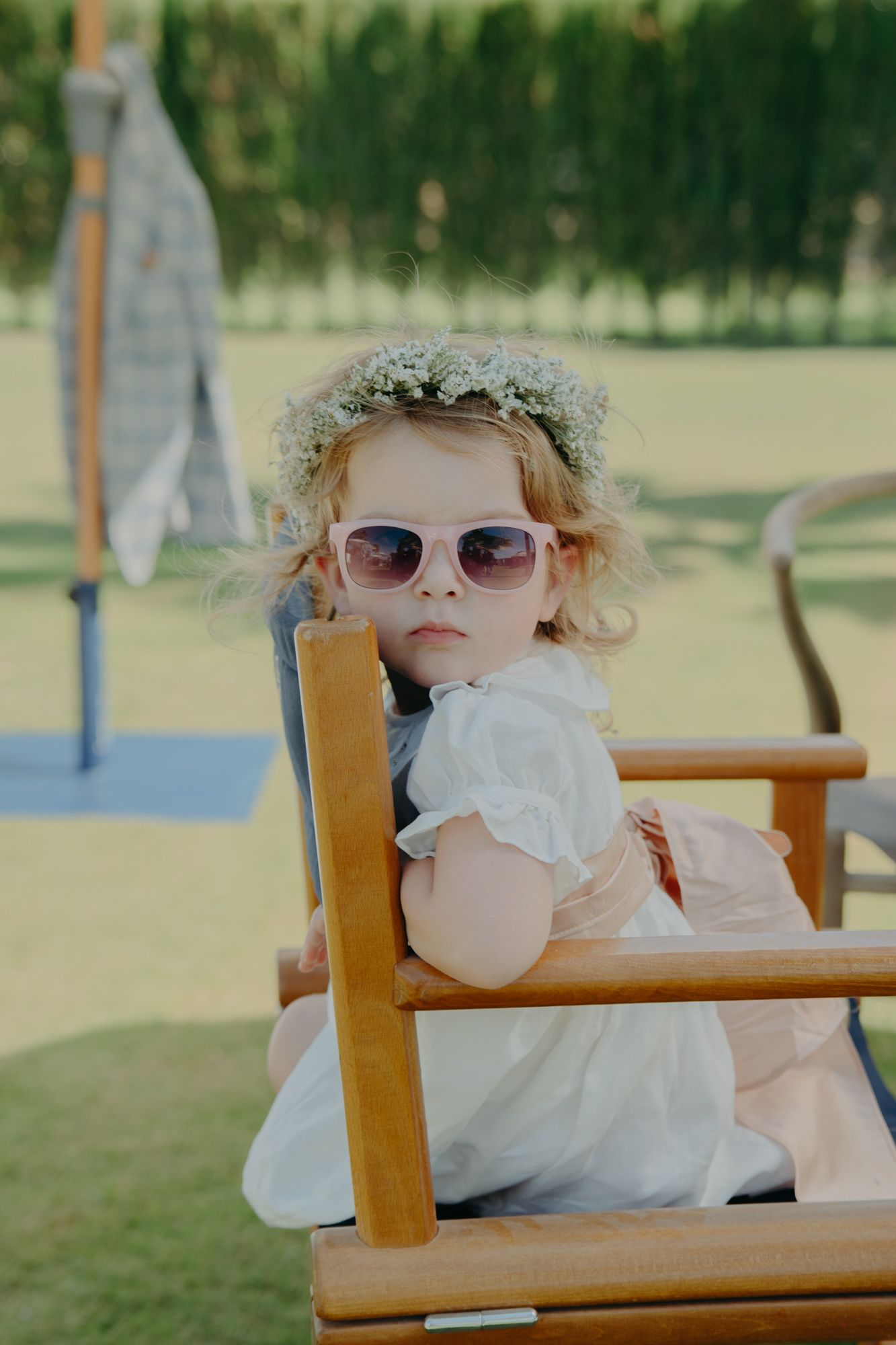 A small figure in a white dress and flower crown sits in a wooden chair on a sunny outdoor lawn.