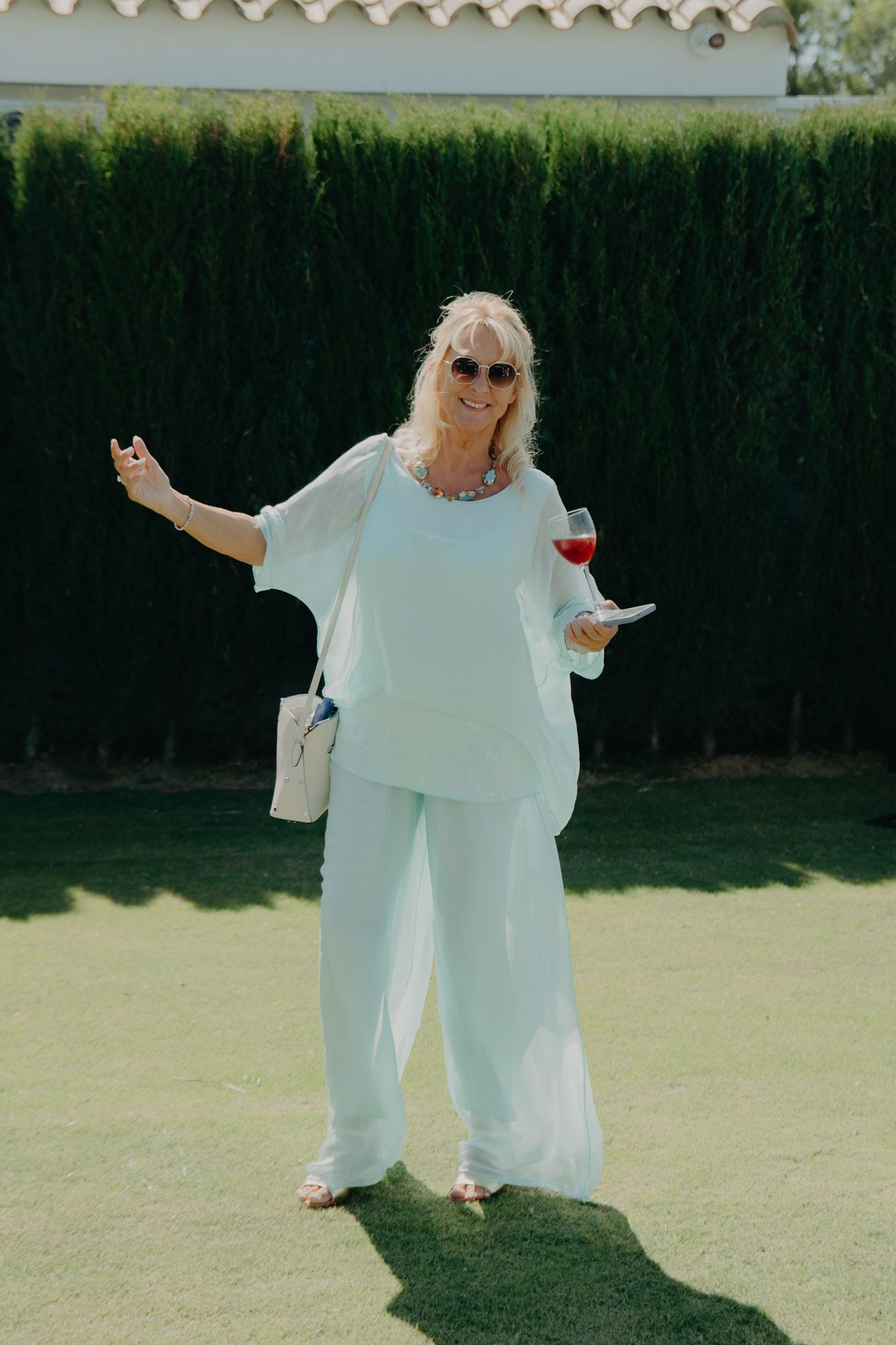 Wedding guest in all-white outfit enjoying drinks on sunny lawn.