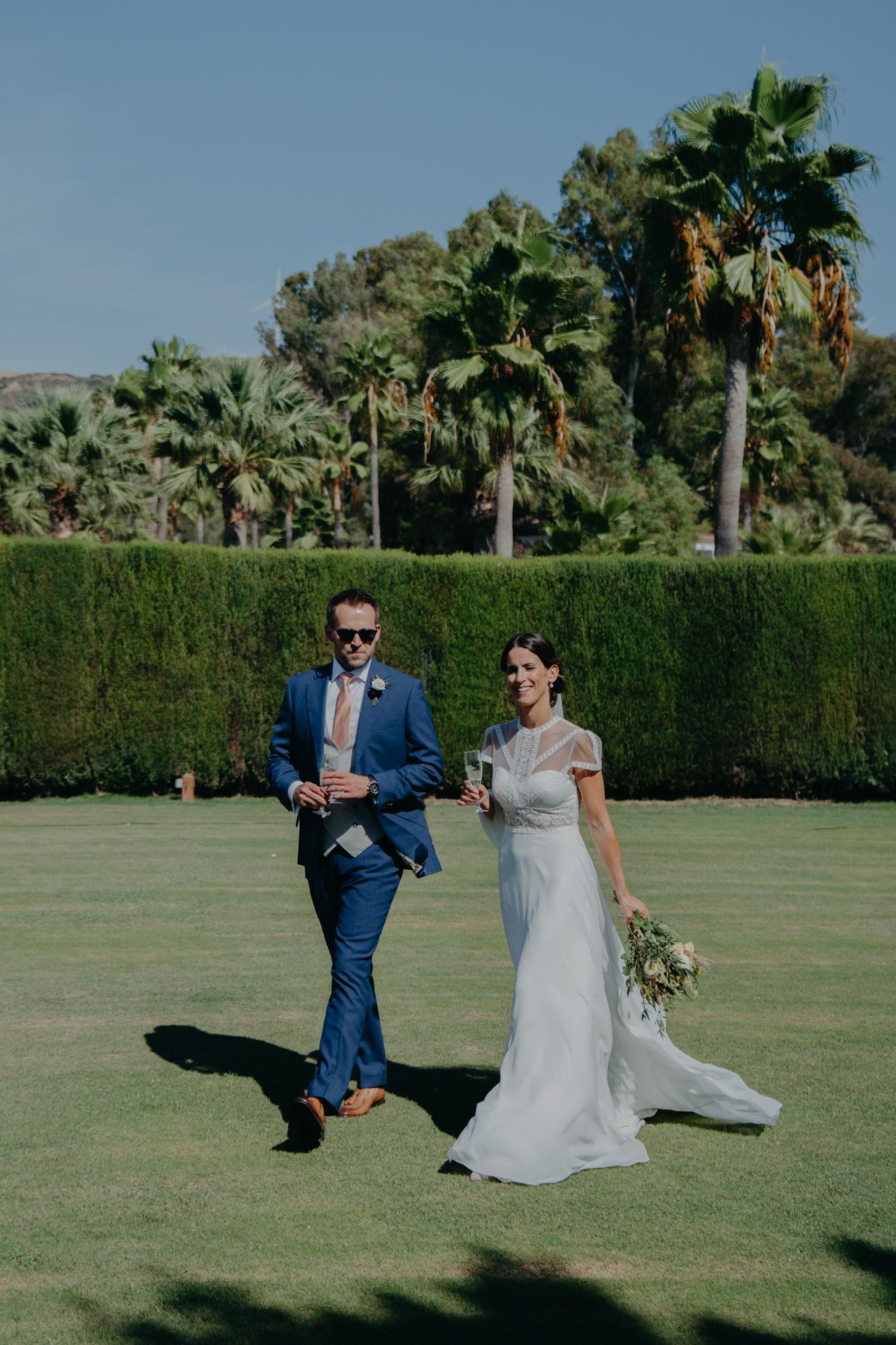 A couple walks across a manicured lawn with palm trees in the background.