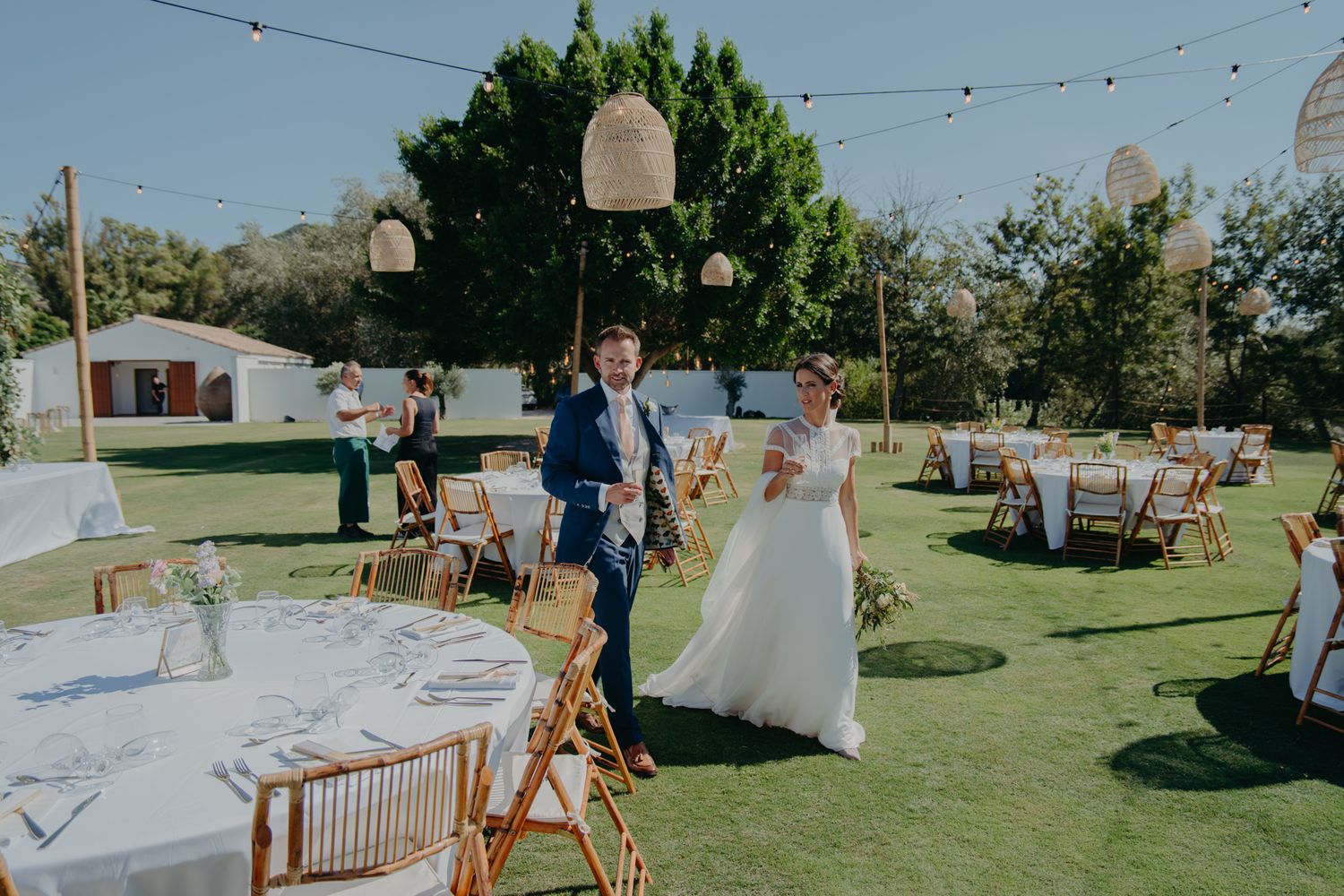 An outdoor wedding reception setup with string lights and tables on a manicured lawn.