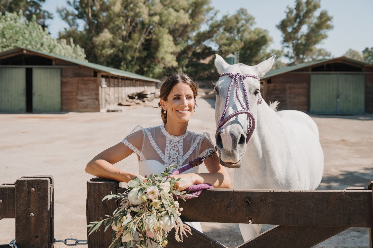 A white horse peeks over a wooden fence as someone holds a beautiful floral bouquet at a rustic barn venue.
