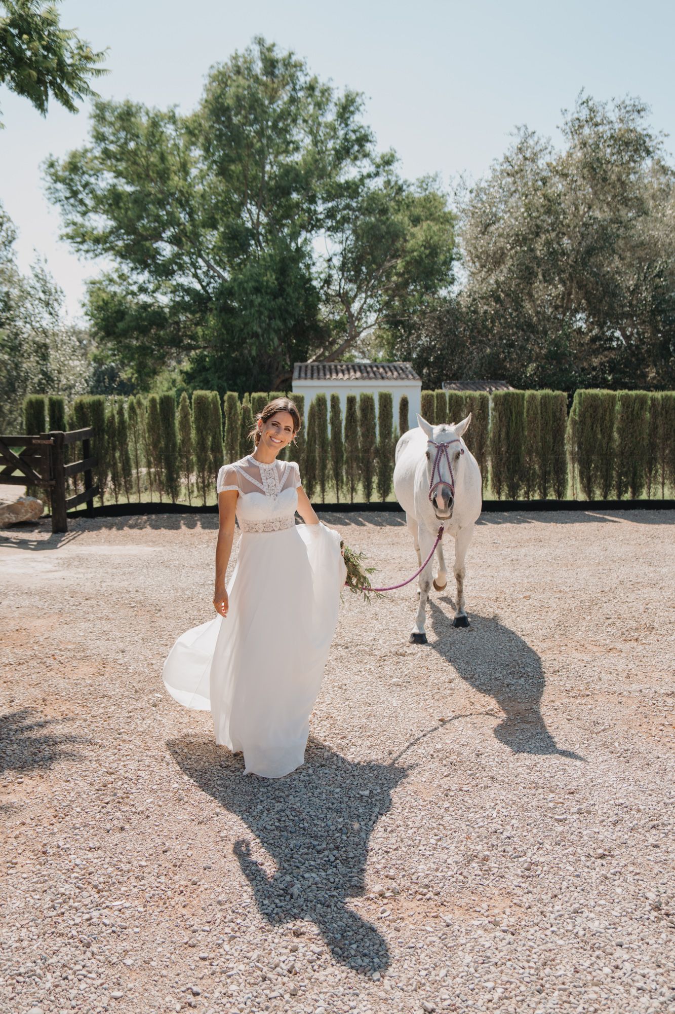 Elegant bride in a flowing white dress poses with a majestic white horse in an outdoor garden setting.