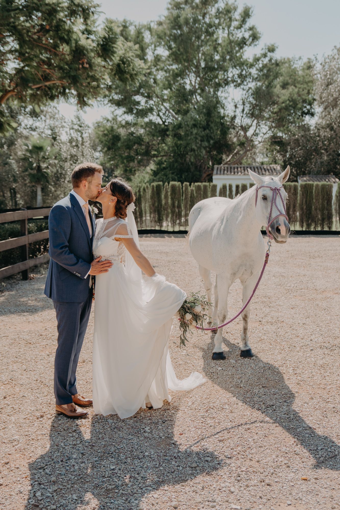Newlyweds share an intimate moment beside a white horse in a rustic outdoor setting.