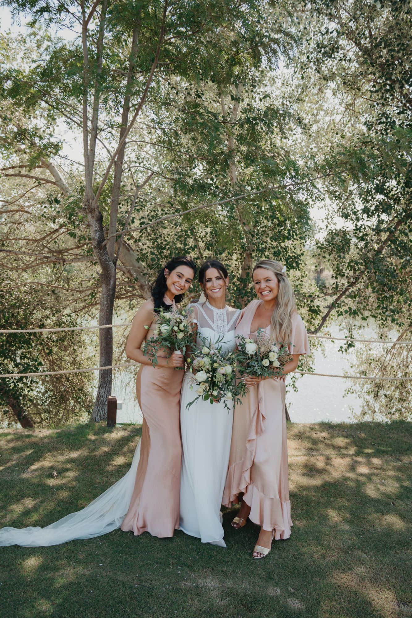Wedding party in blush pink dresses poses outdoors under green trees with floral bouquets.