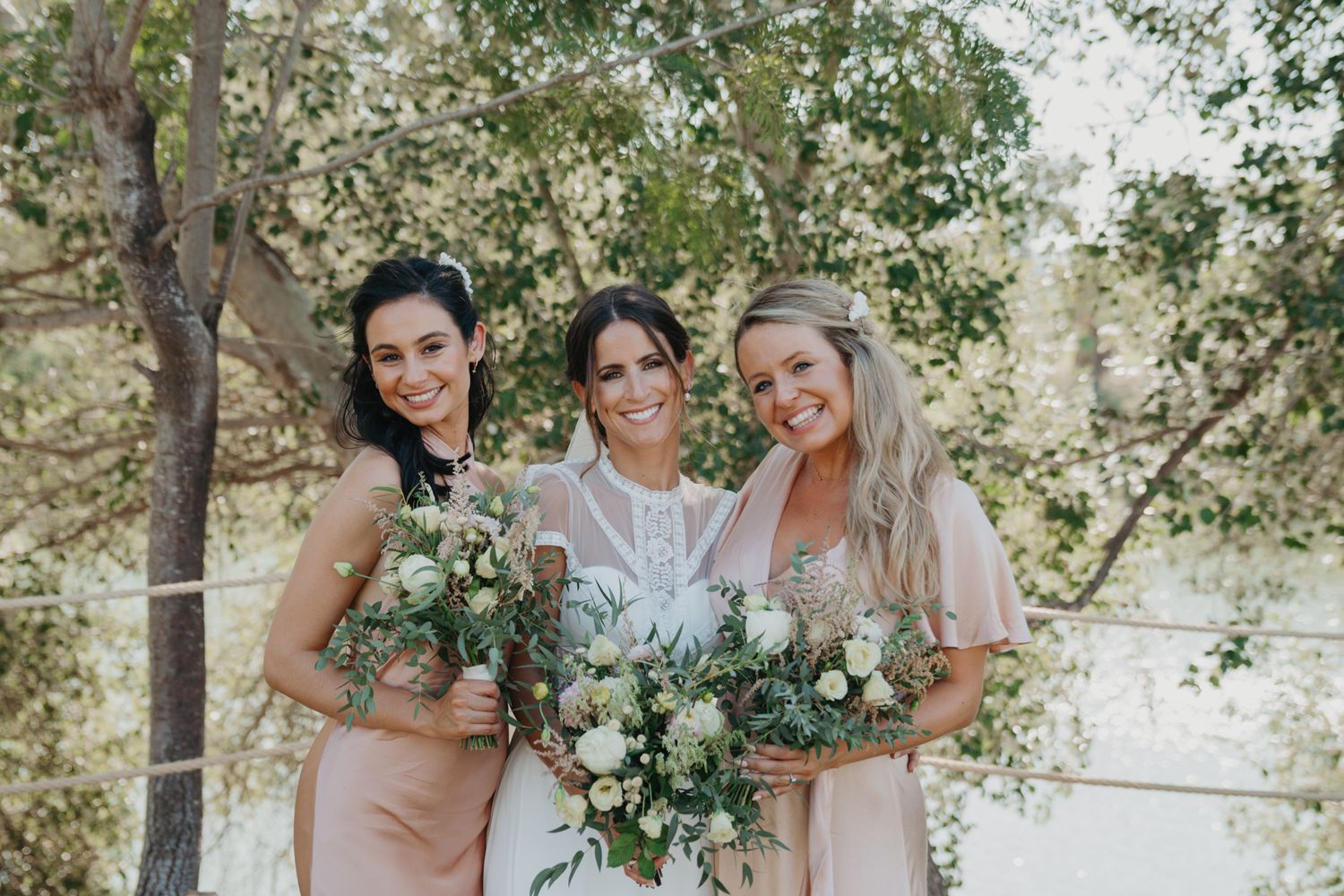 Three attendants holding elegant floral bouquets pose outdoors with natural greenery.