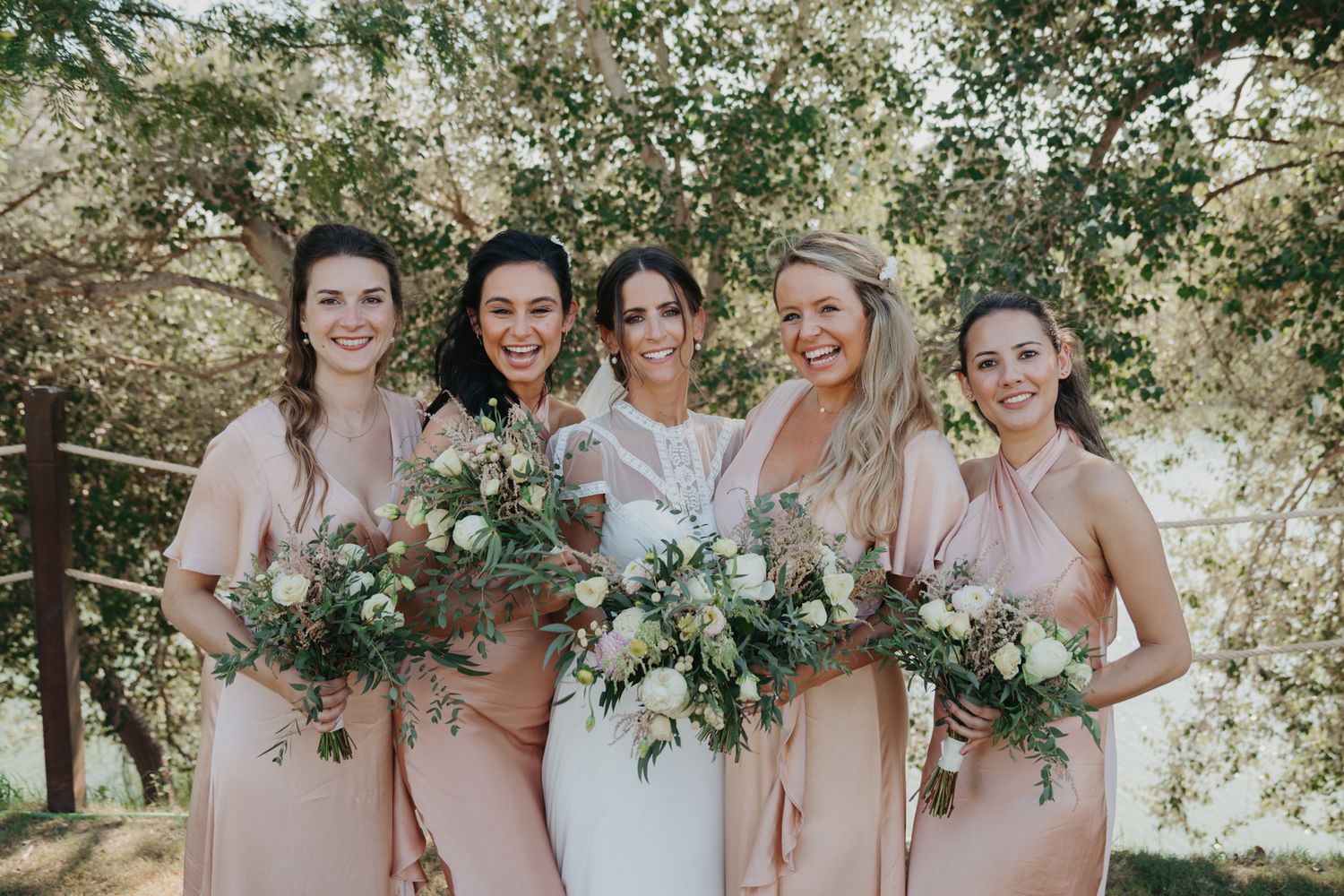 Wedding party in blush pink dresses holding fresh green and white bouquets outdoors.