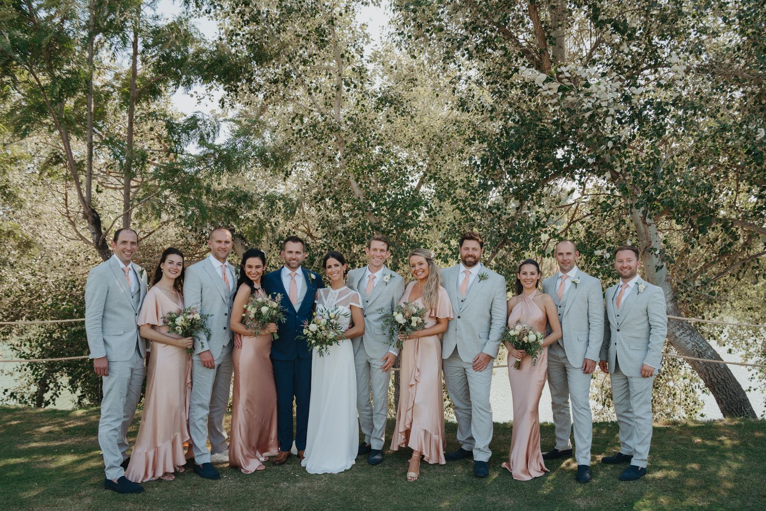 Wedding party in pastel pink and gray attire poses under trees for group photo.