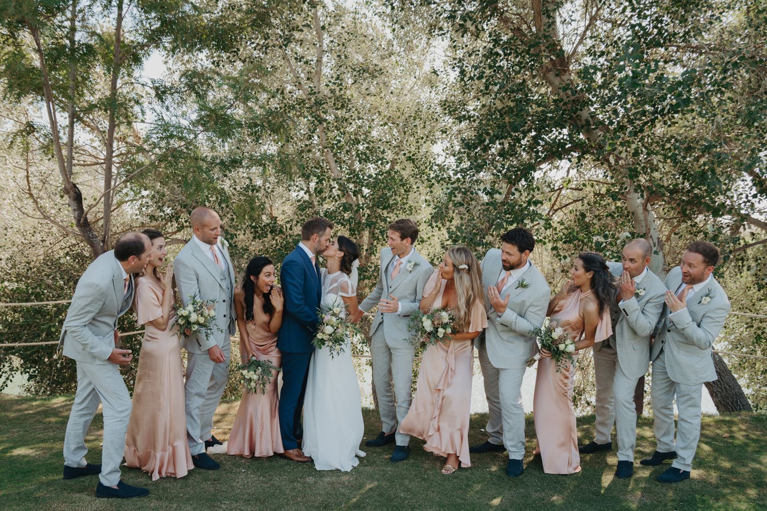 Wedding party in pastels and light colors posed together outdoors under trees.