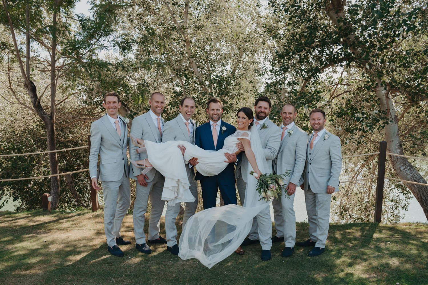 Wedding party in light gray suits and a navy blue suit pose together outdoors on a sunny day amongst trees.