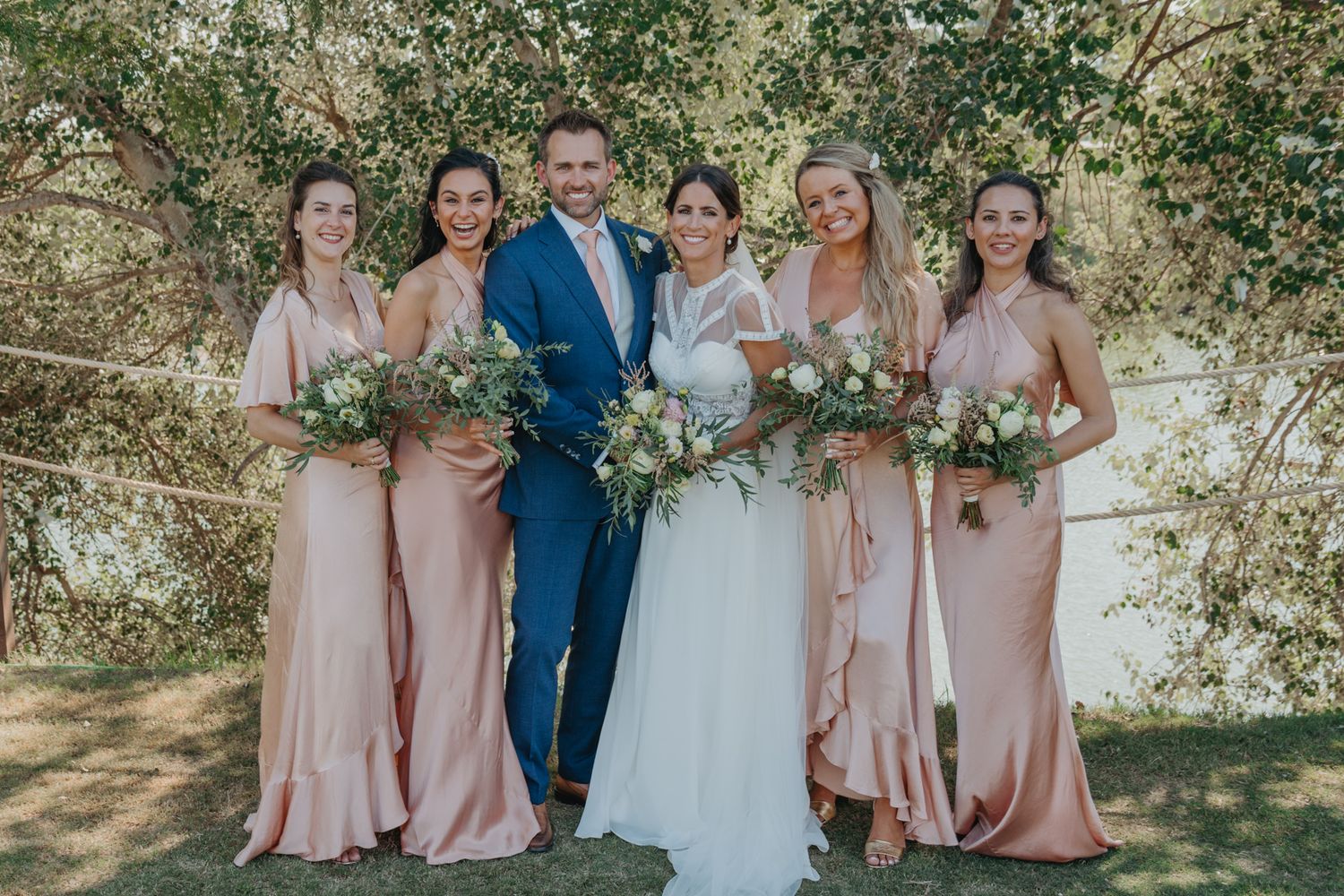 Wedding party poses outdoors wearing blush pink dresses with floral bouquets.