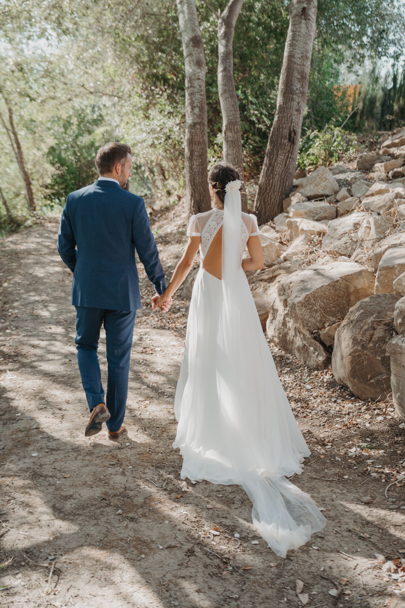 Couple walking hand in hand on a rocky path, bride's white dress flowing behind them.