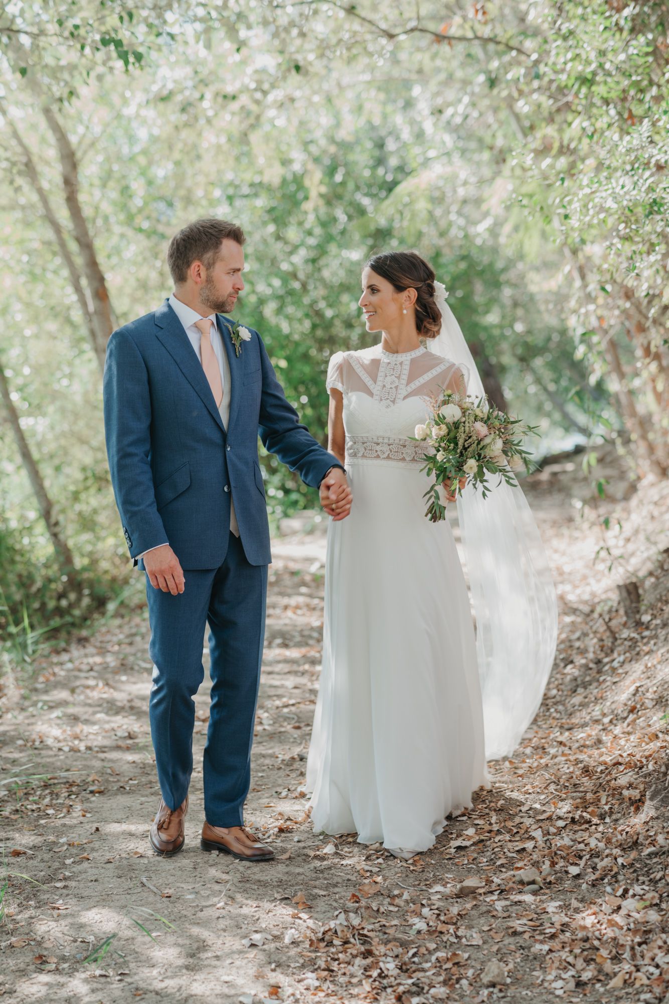 A romantic wedding couple walking on a wooded path, with the bride in white and groom in navy blue.
