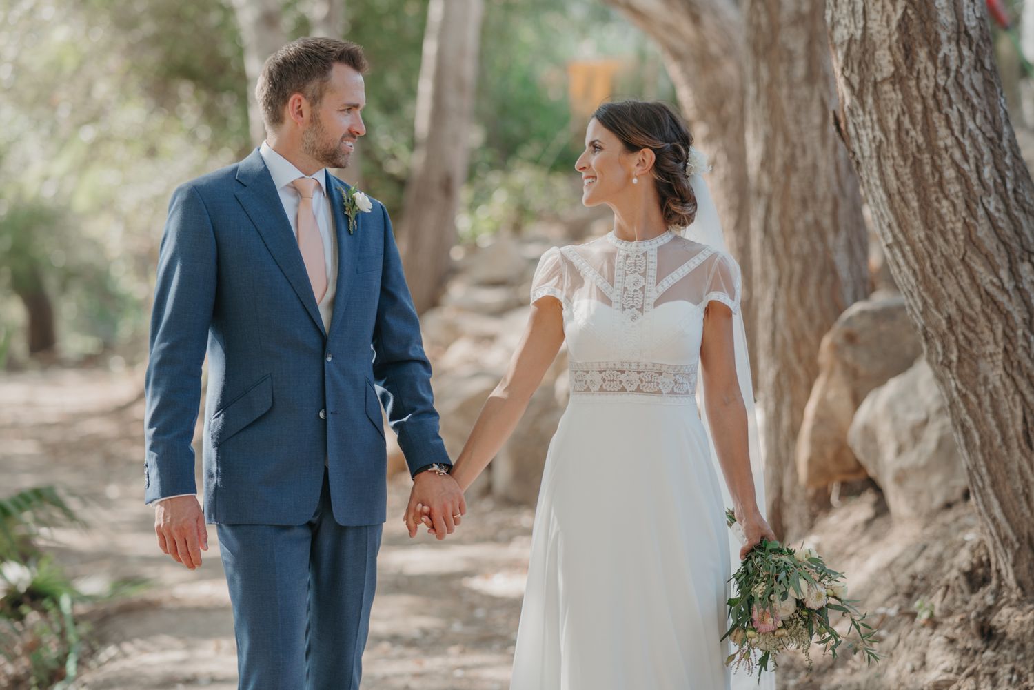 Couple walks hand in hand along a tree-lined path in wedding attire.