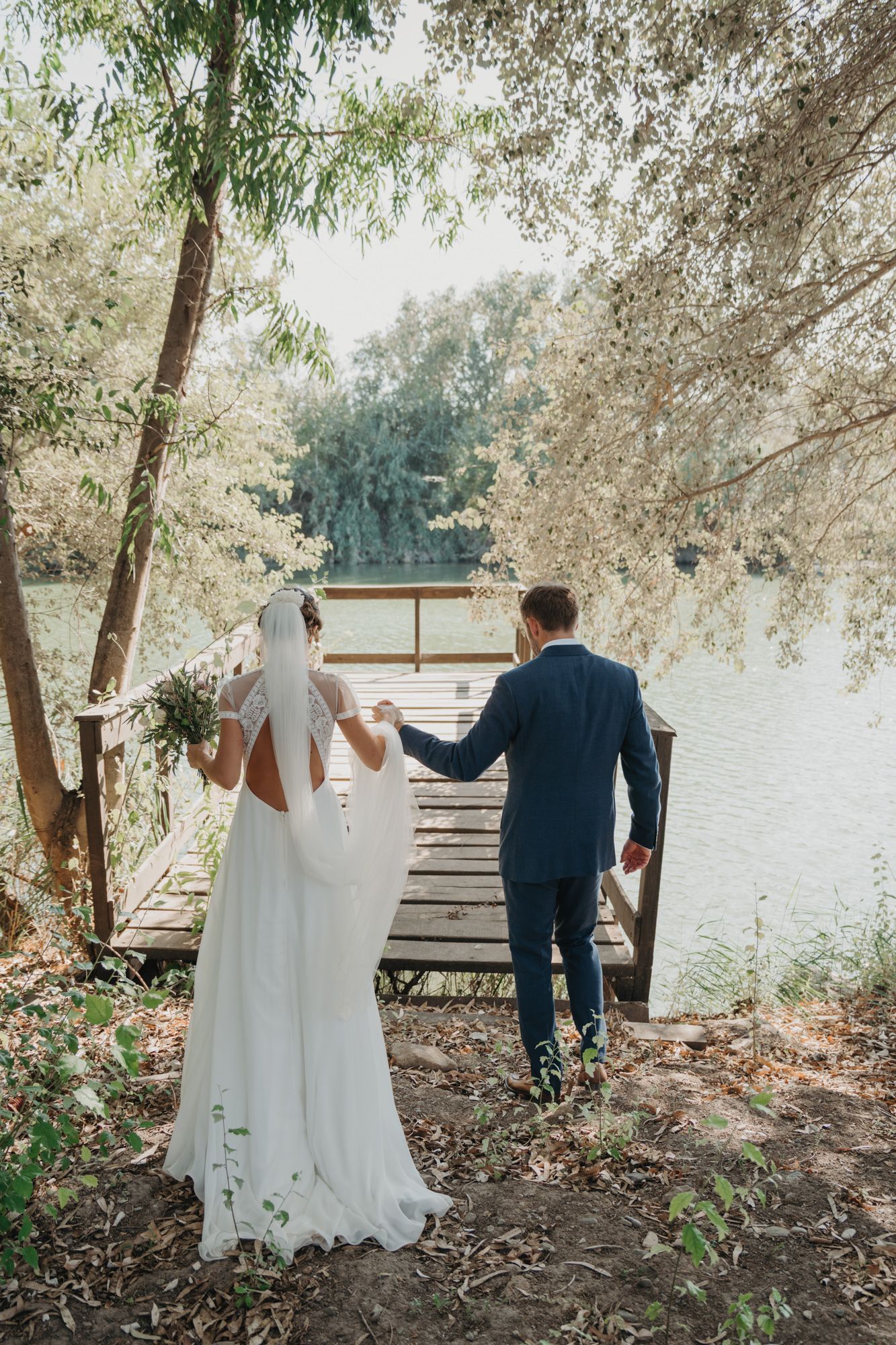 A wedding couple stands together on a wooden dock overlooking a serene lake surrounded by trees.