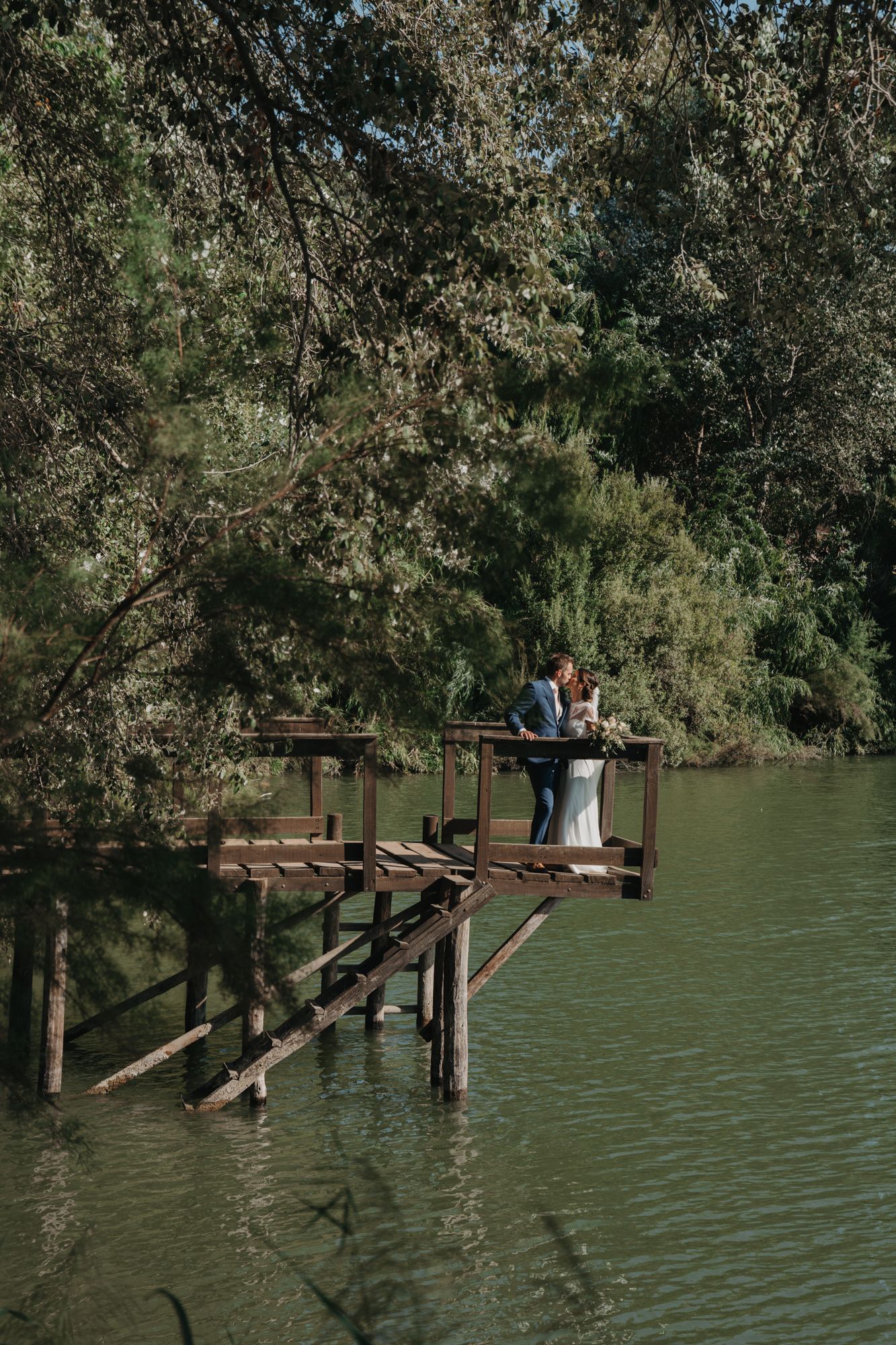 A couple stands together on a wooden dock overlooking a serene lake surrounded by trees.