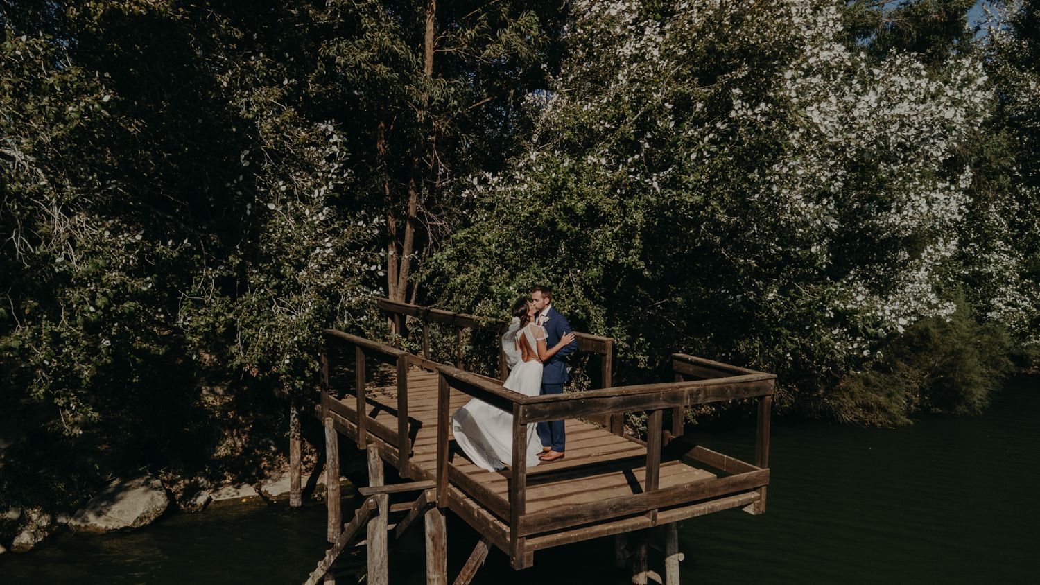 A scenic lakeside wooden deck surrounded by trees creates a romantic wedding photo setting.