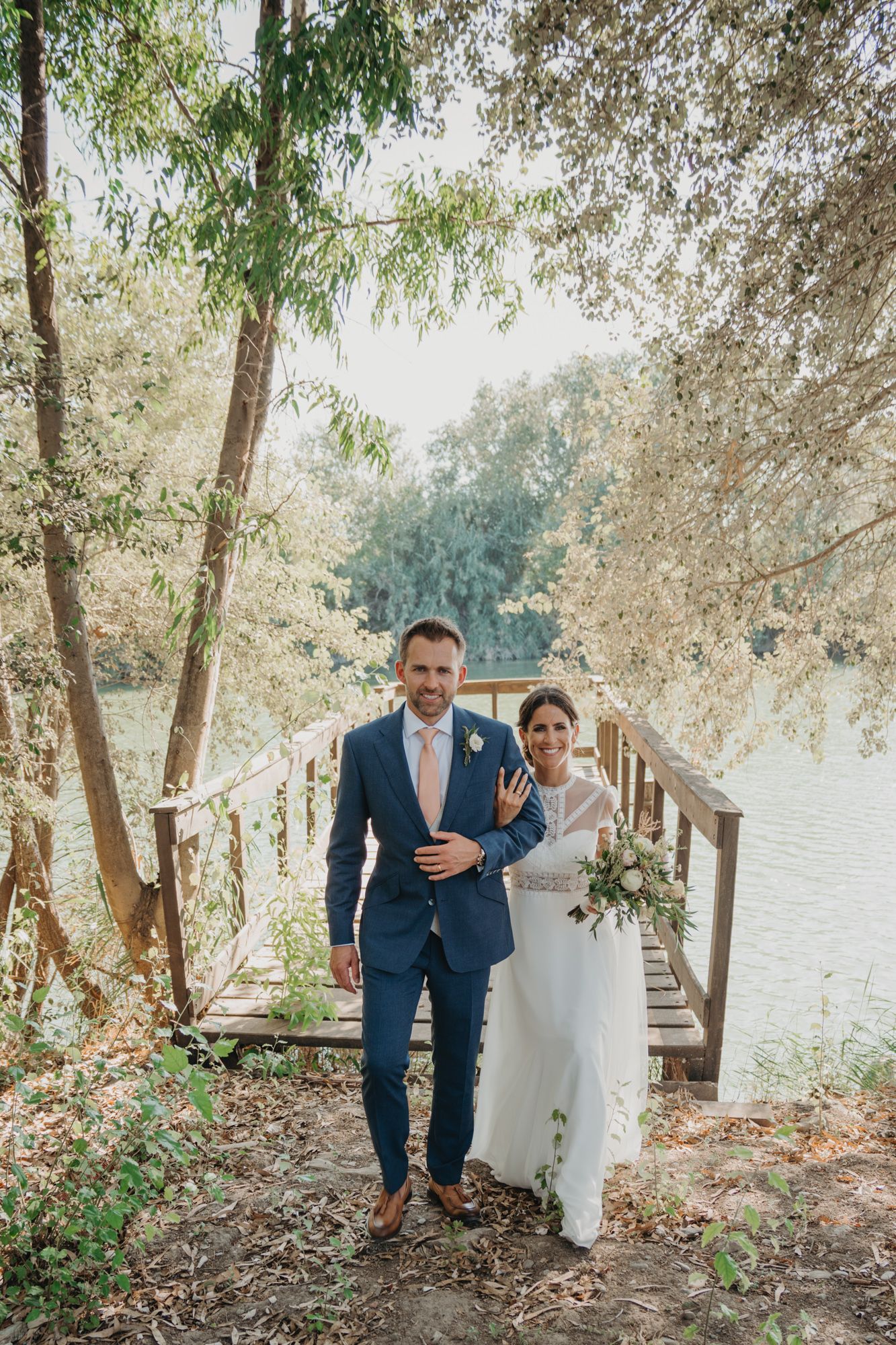 A wedding photoshoot on a rustic wooden bridge surrounded by willow trees near a tranquil water body.