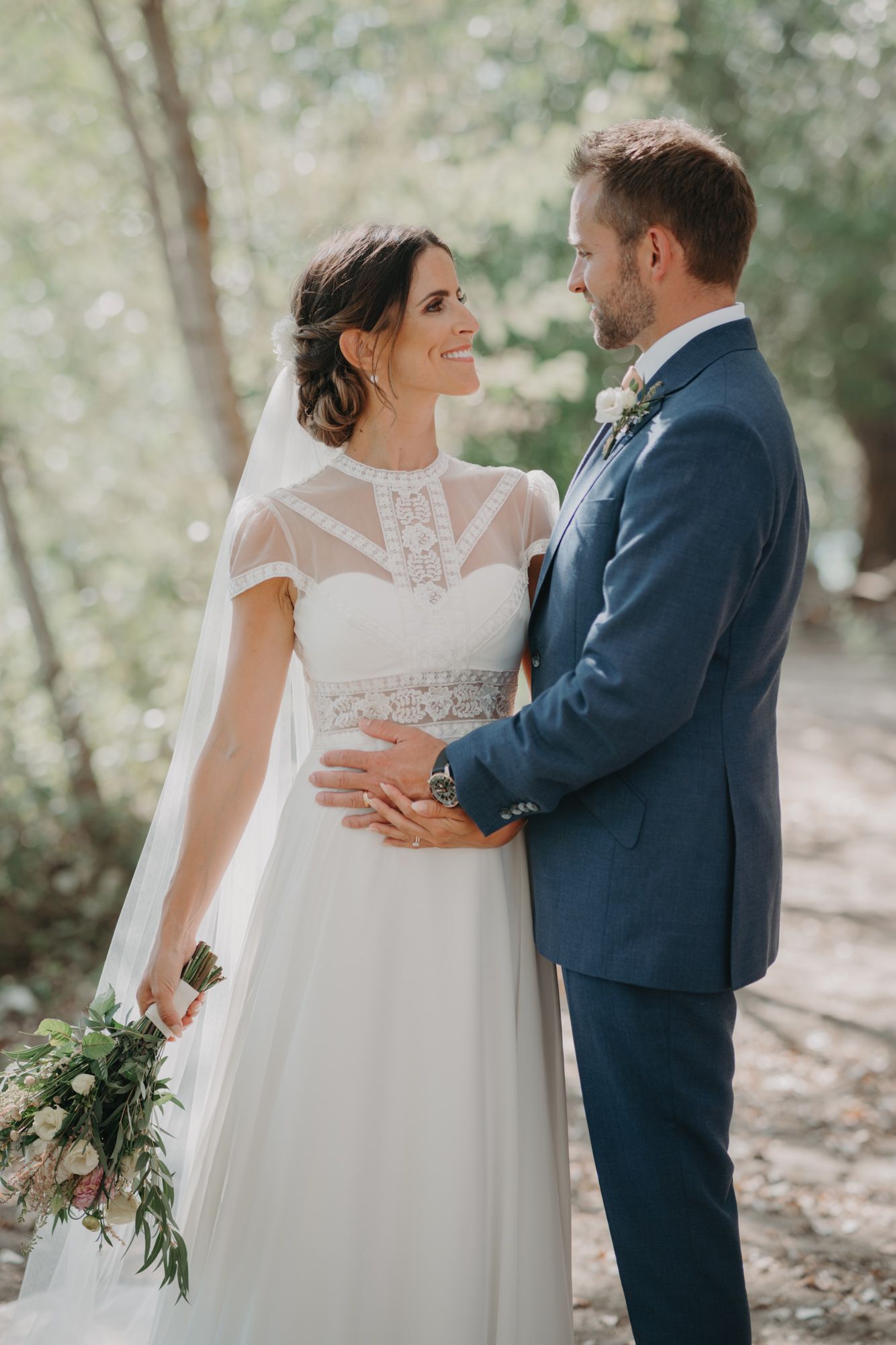 A romantic wedding couple shares a tender moment in a wooded setting with soft natural lighting.
