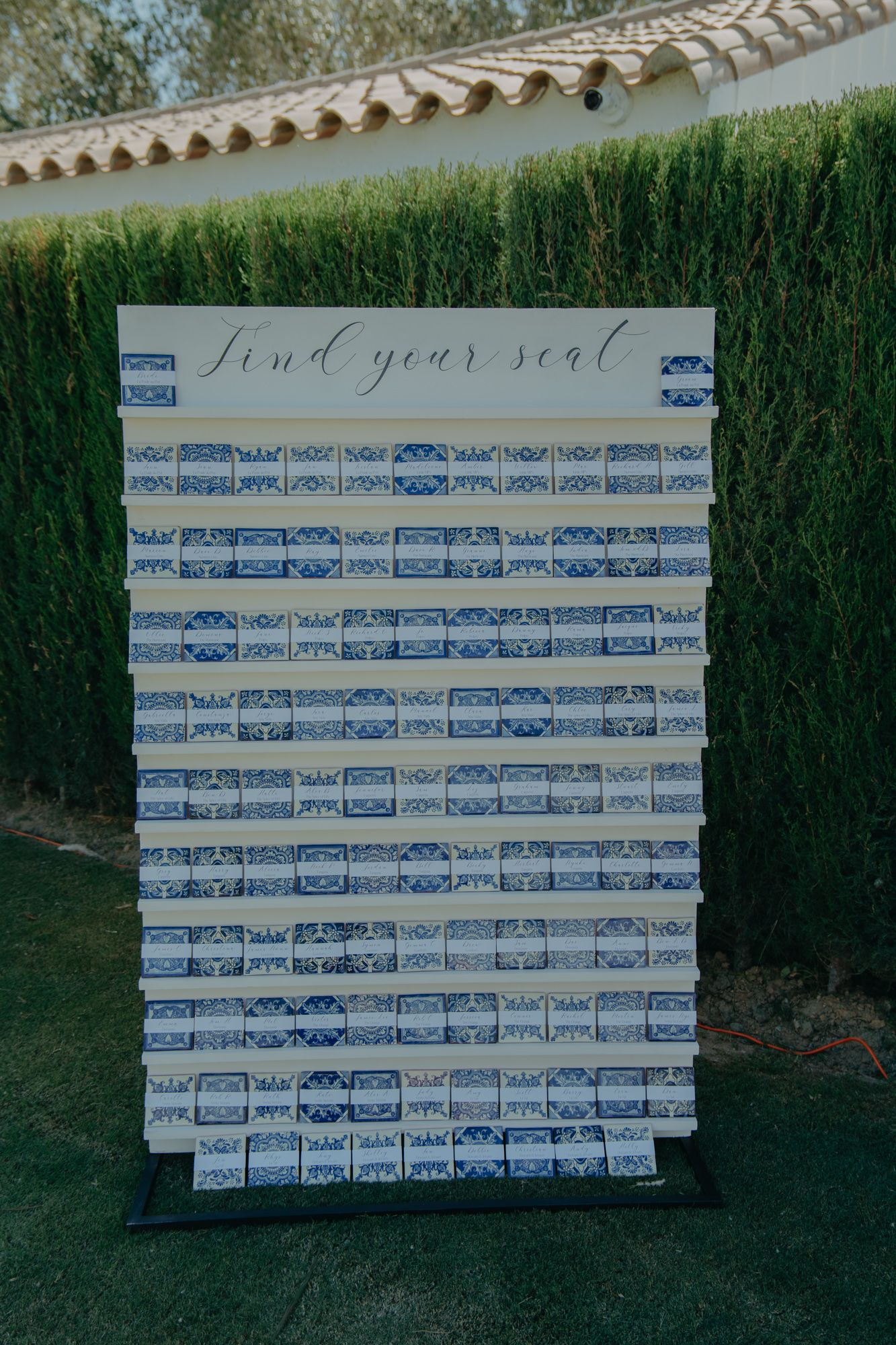 A wedding seating chart display with blue cards arranged on a white board against a hedge wall.