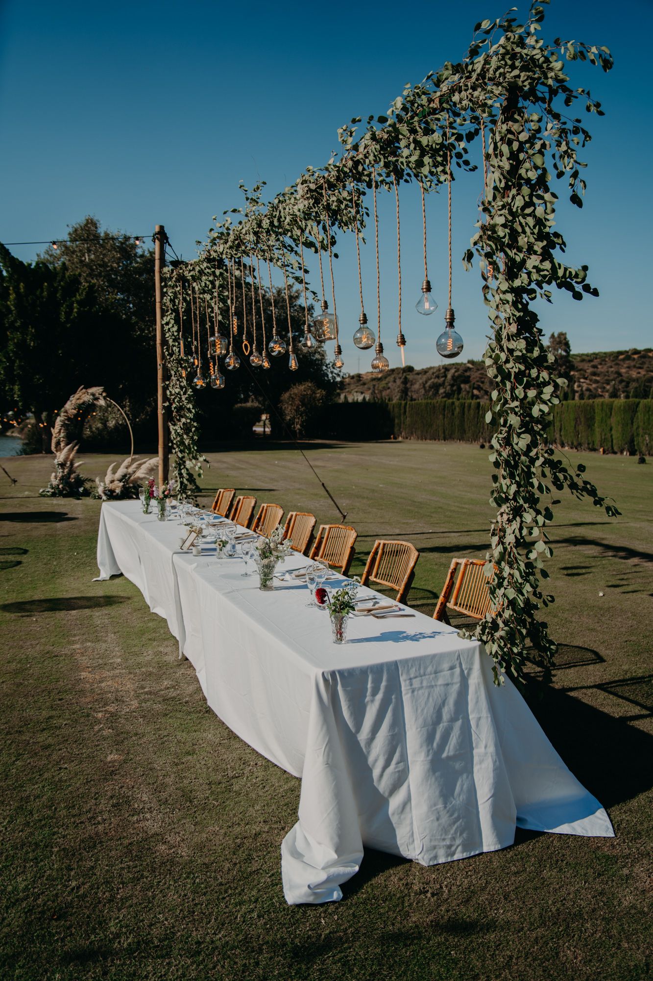An elegant outdoor table setting with hanging Edison bulbs and greenery arch for a wedding reception.