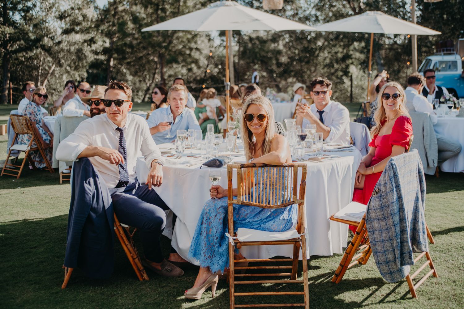 Wedding guests enjoying an outdoor reception with white umbrellas and table settings on a sunny lawn.