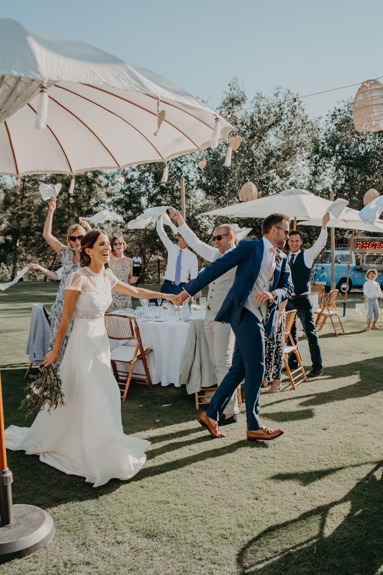 Newlyweds and wedding party celebrating outdoors under white umbrellas on a sunny day.