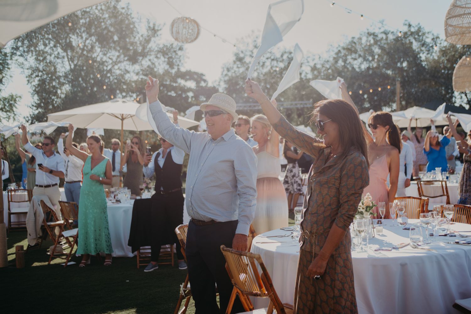 Wedding celebration on a lawn with umbrellas and guests toasting amid sunlit garden party atmosphere.