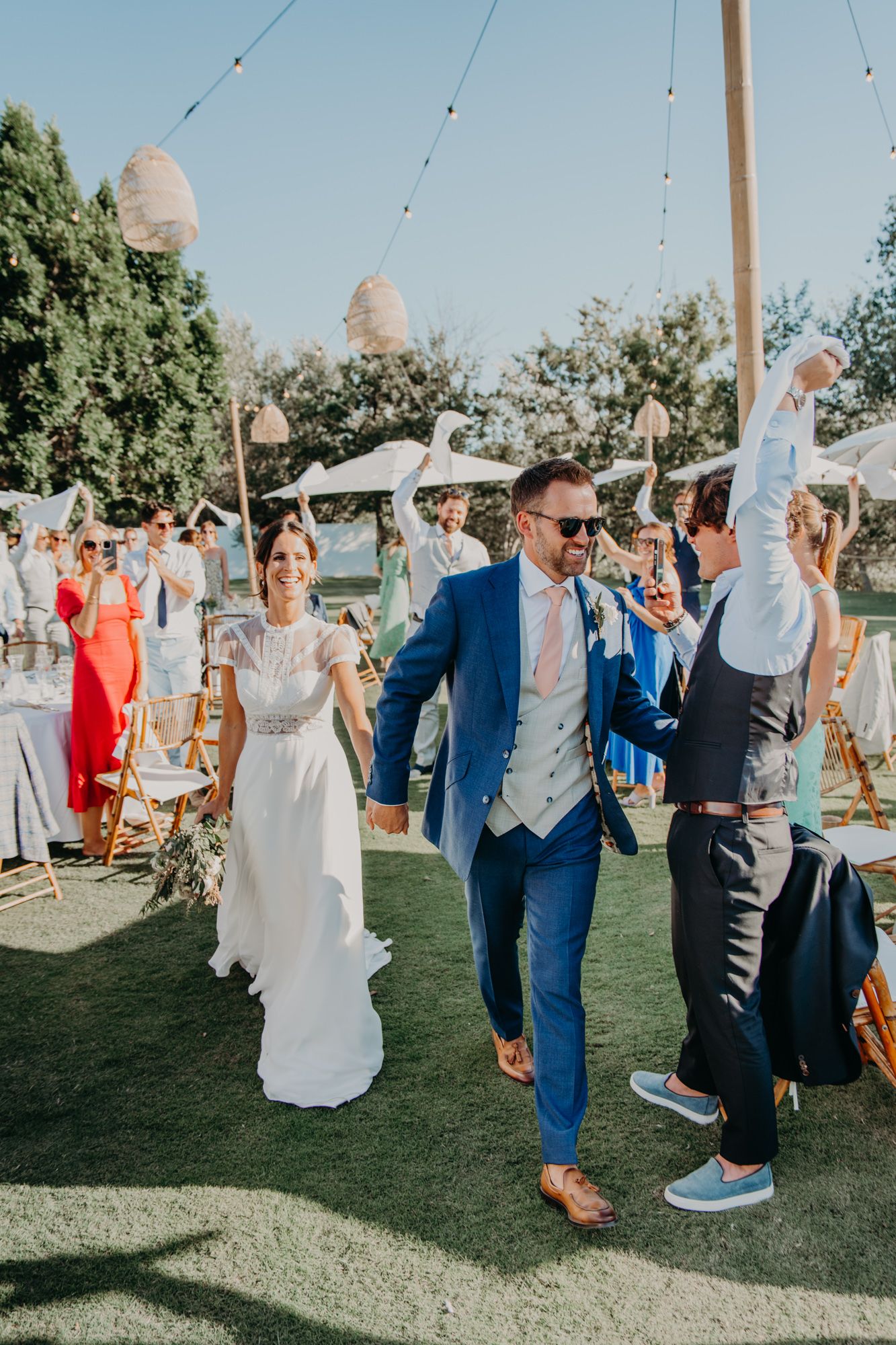 Outdoor daytime wedding celebration with guests dancing on a lawn under string lights.