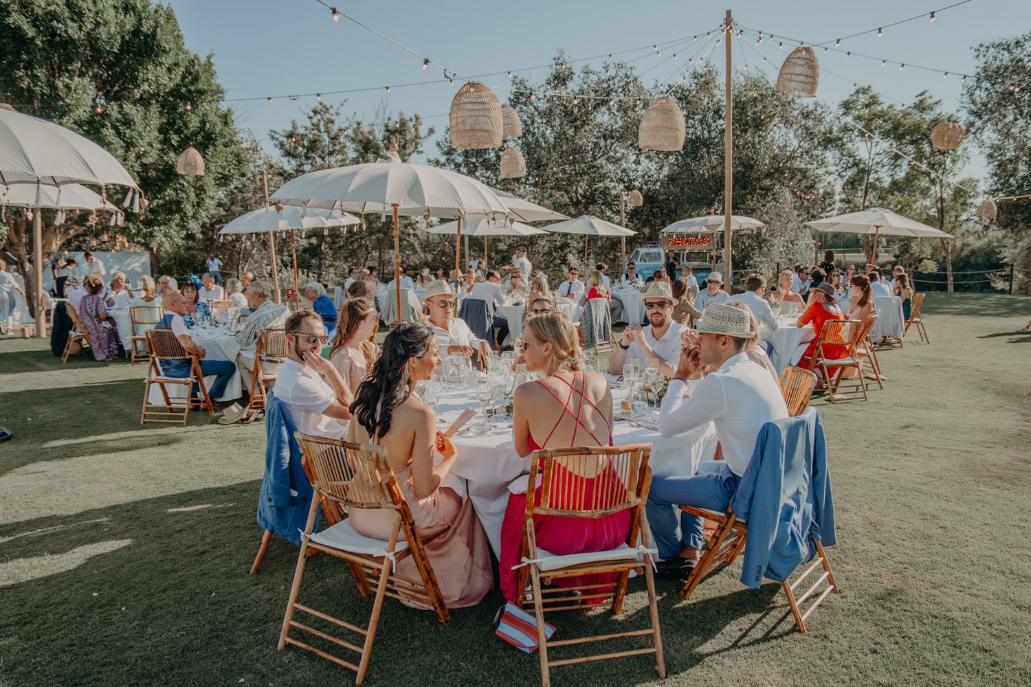 Outdoor garden wedding reception with round tables, white umbrellas and string lights decorating a sunny lawn space.