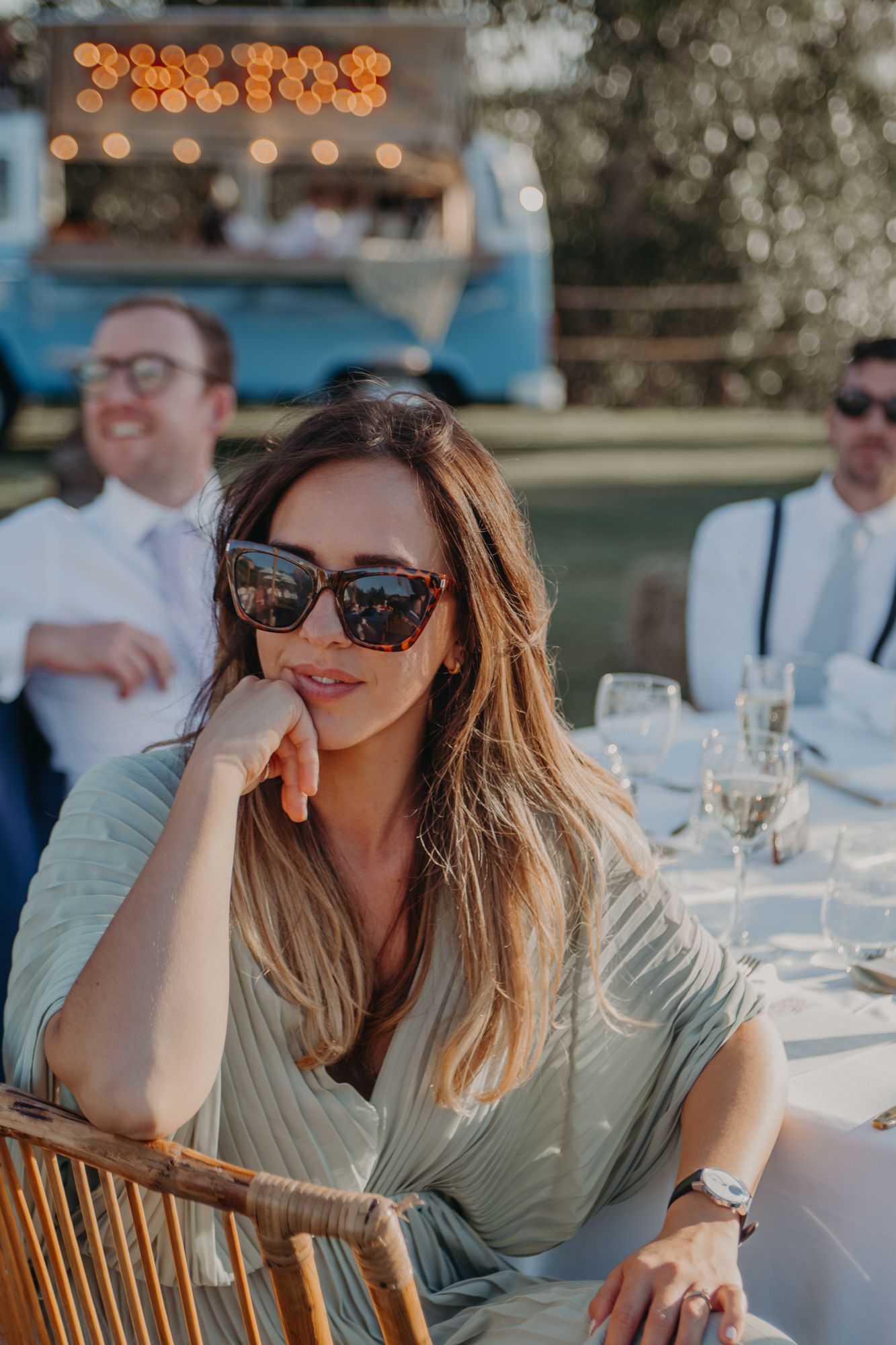 Guest in sunglasses relaxing at outdoor wedding reception with string lights in background.