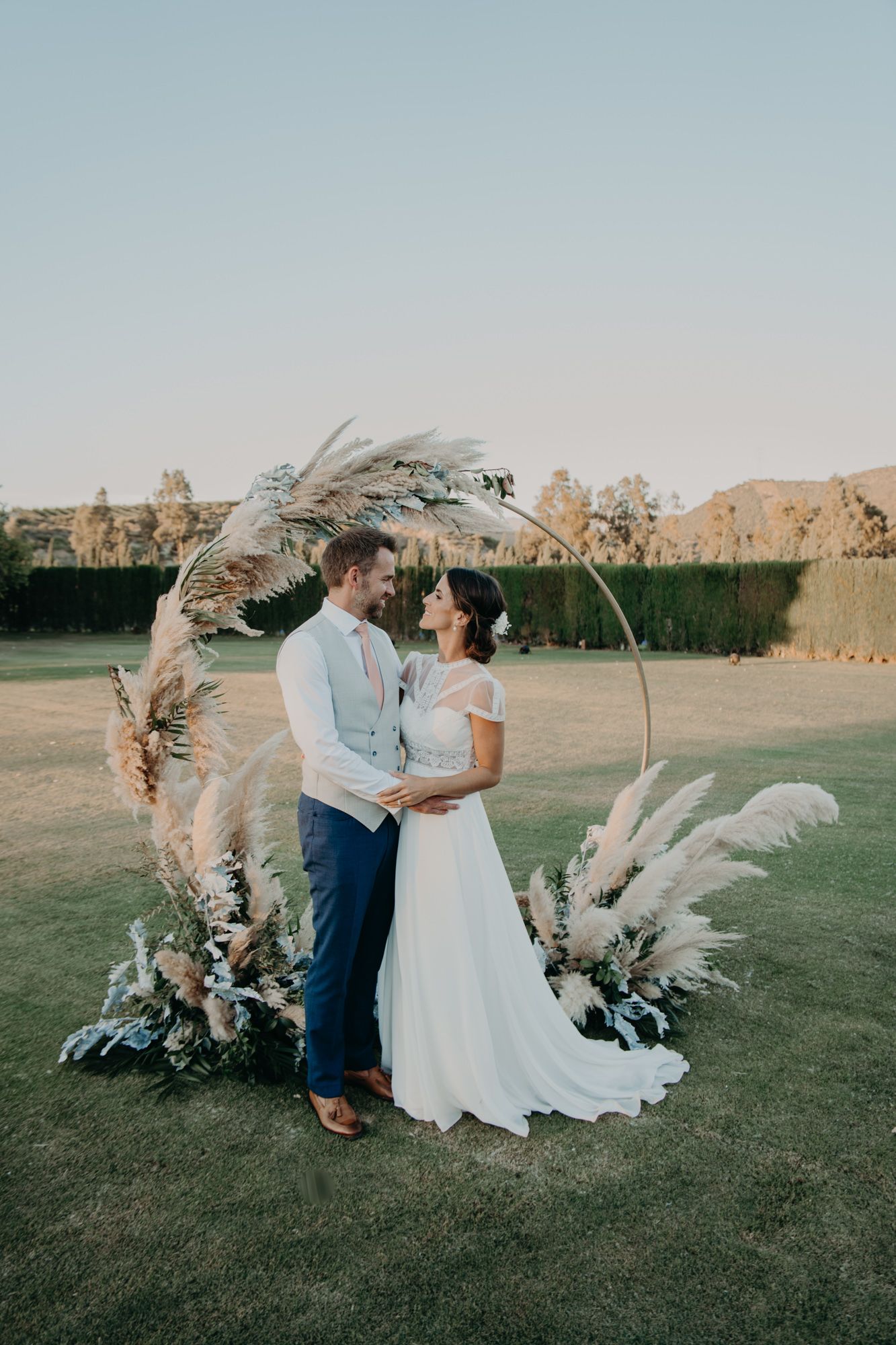A wedding couple stands beneath an elegant curved arch decorated with pampas grass and dried flowers.