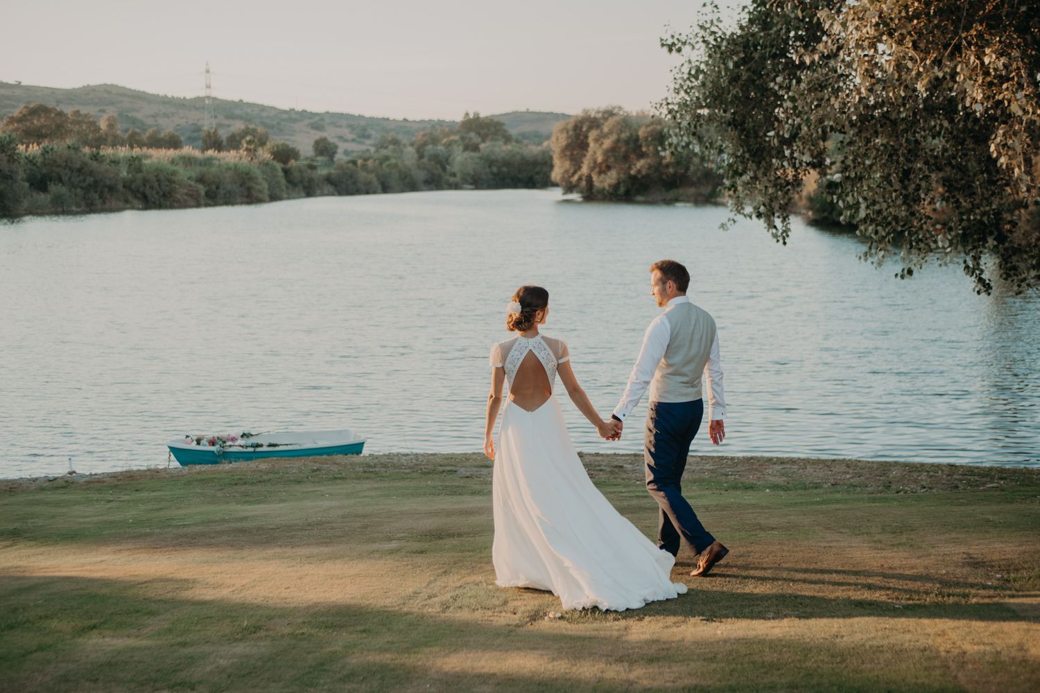 Newlyweds walk hand in hand along a lakeside path with a small boat in the background at sunset.