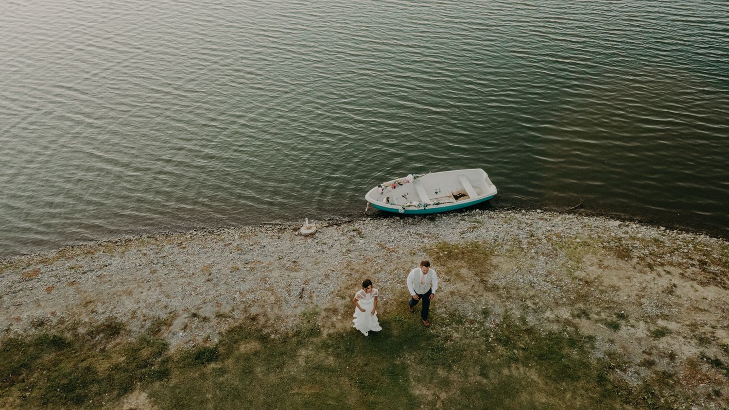 An aerial view of a couple standing on a cliff edge near a small boat on calm waters.