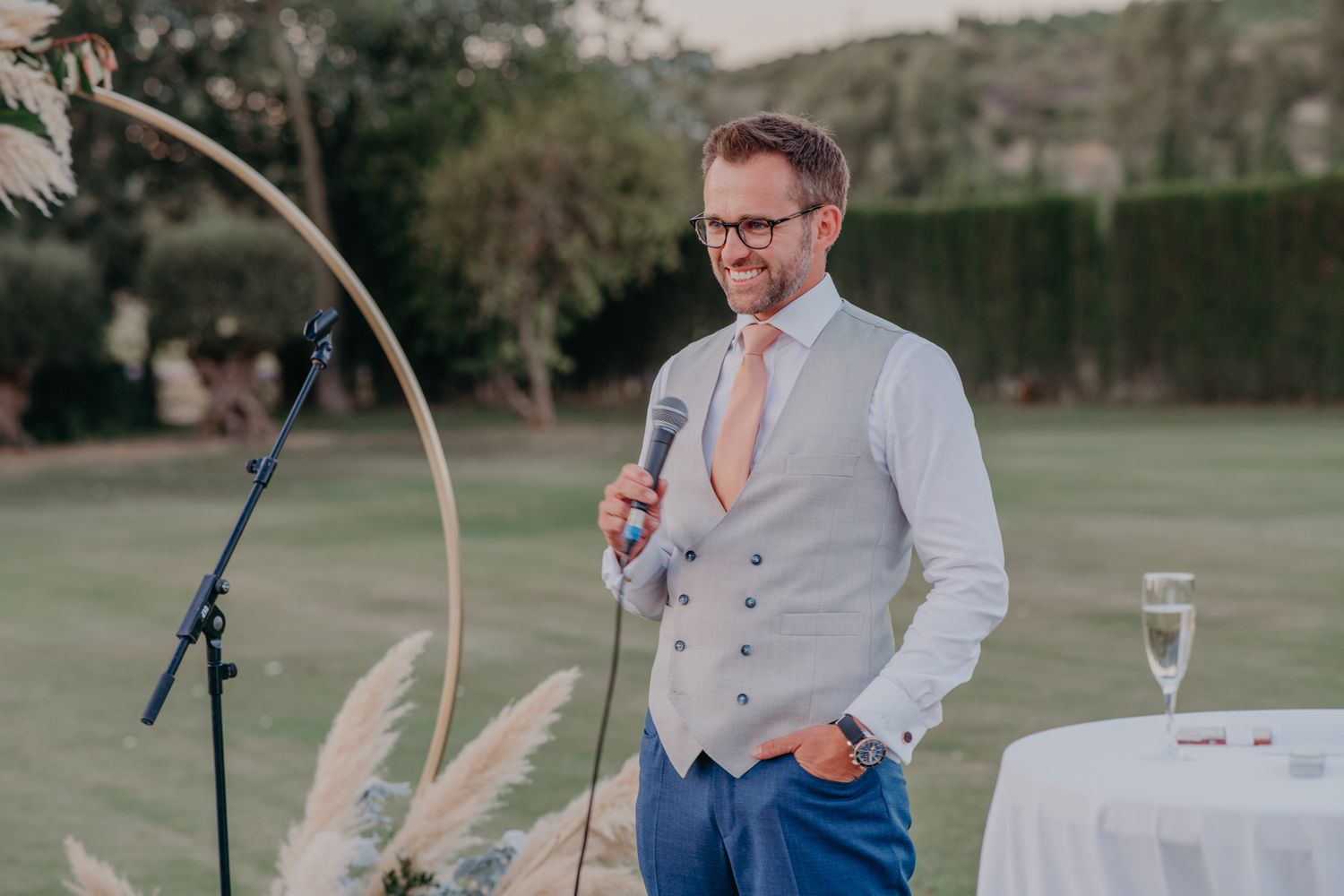 A person speaks at a microphone during an outdoor wedding reception while dressed in a vest and tie.