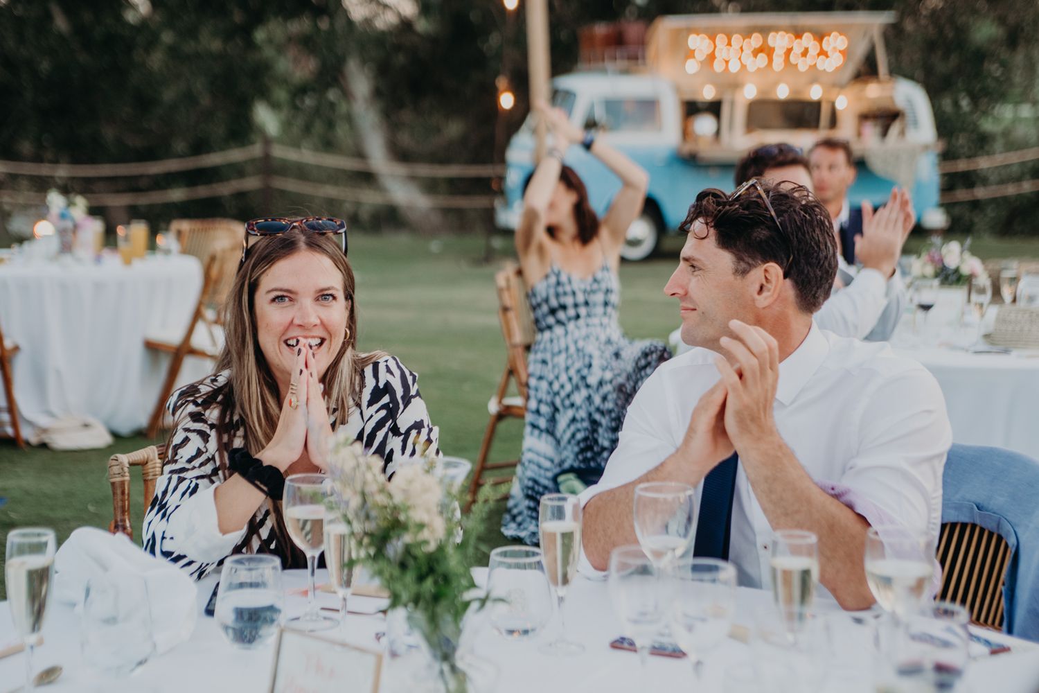 Wedding reception guests react with joy and surprise during an outdoor dinner celebration.