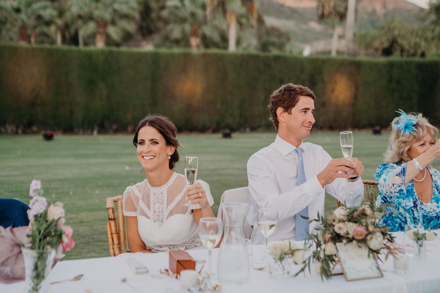 Wedding guests seated at an elegant outdoor reception table with floral centerpieces during sunset.