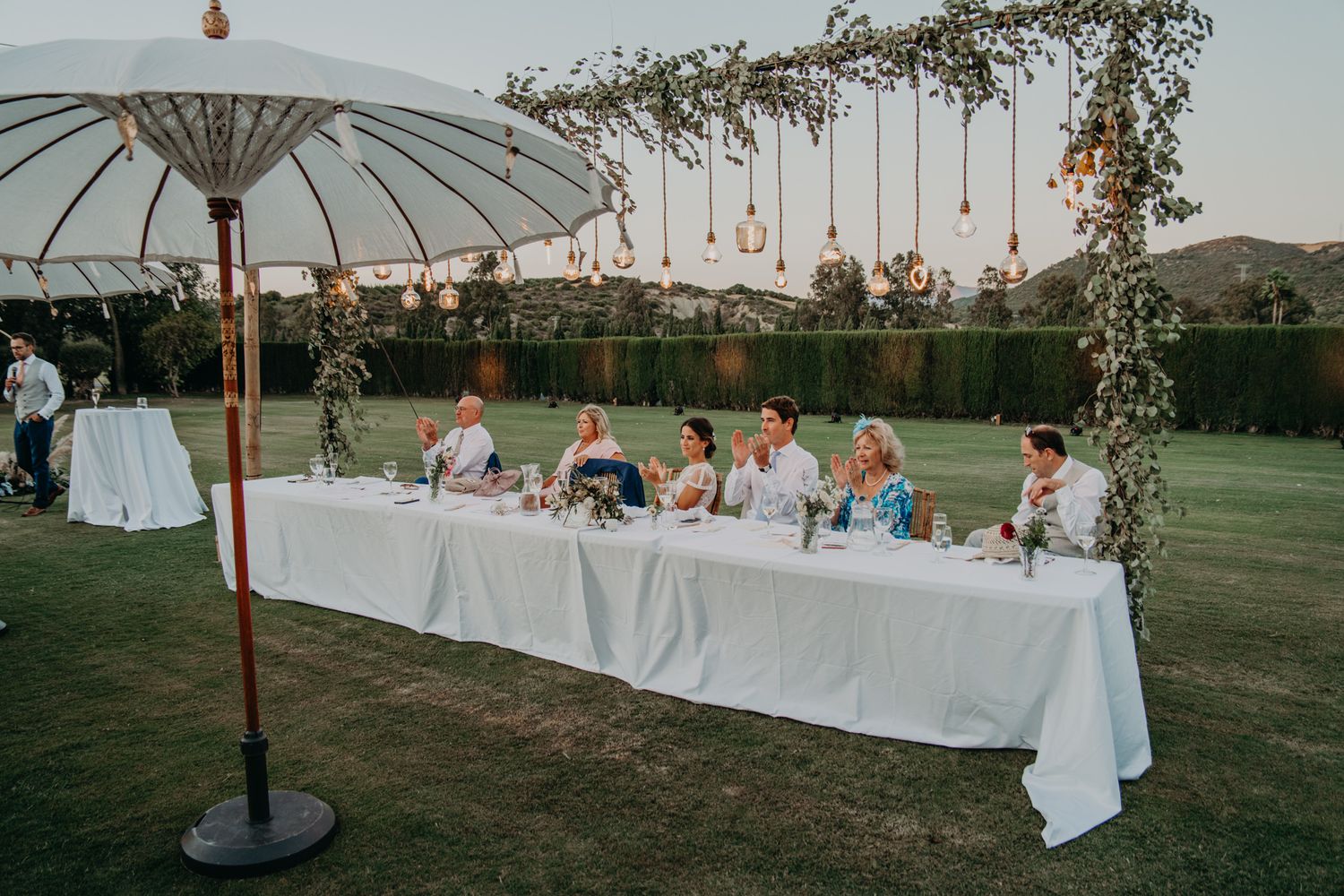 Outdoor wedding reception with a long white table under hanging lights and white parasol.
