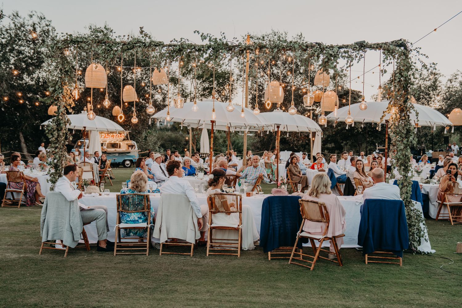 An outdoor wedding reception features elegant chandeliers hanging over tables set for dining at dusk.