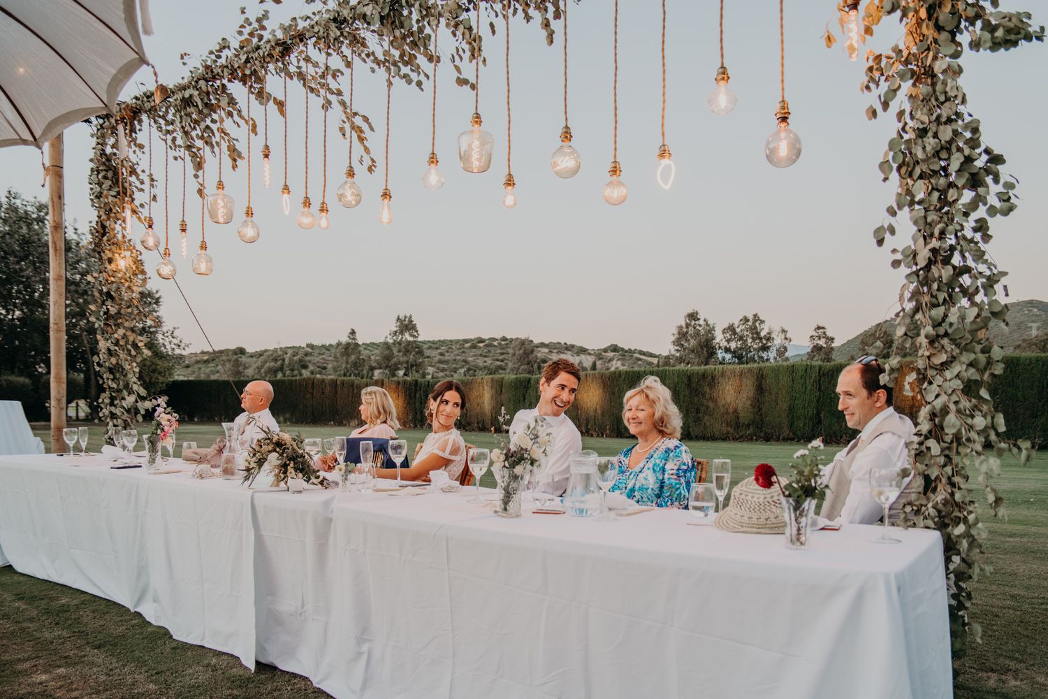 Wedding reception dining table decorated with hanging lights and floral arrangements at sunset.