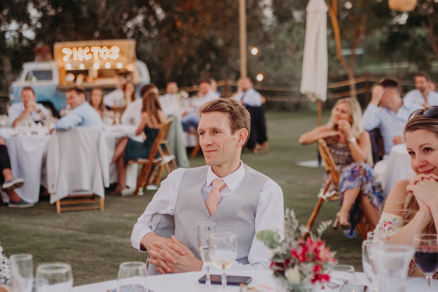 A wedding reception held outdoors with guests seated at tables and a pink flower centerpiece in the foreground.