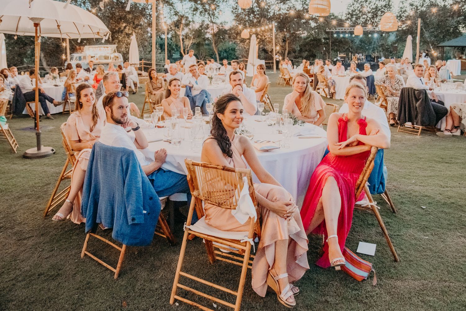 Guests seated at outdoor reception tables on grass lawn with decorative lighting at sunset celebration event.