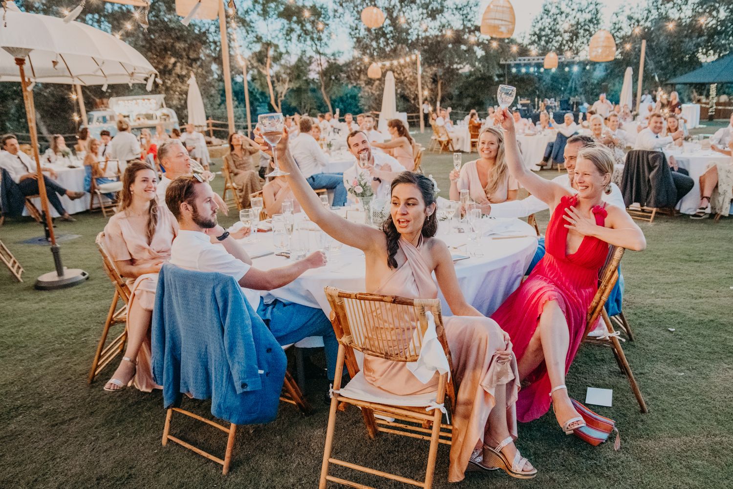 Outdoor wedding reception guests seated at tables on a lawn with string lights and umbrellas during golden hour.