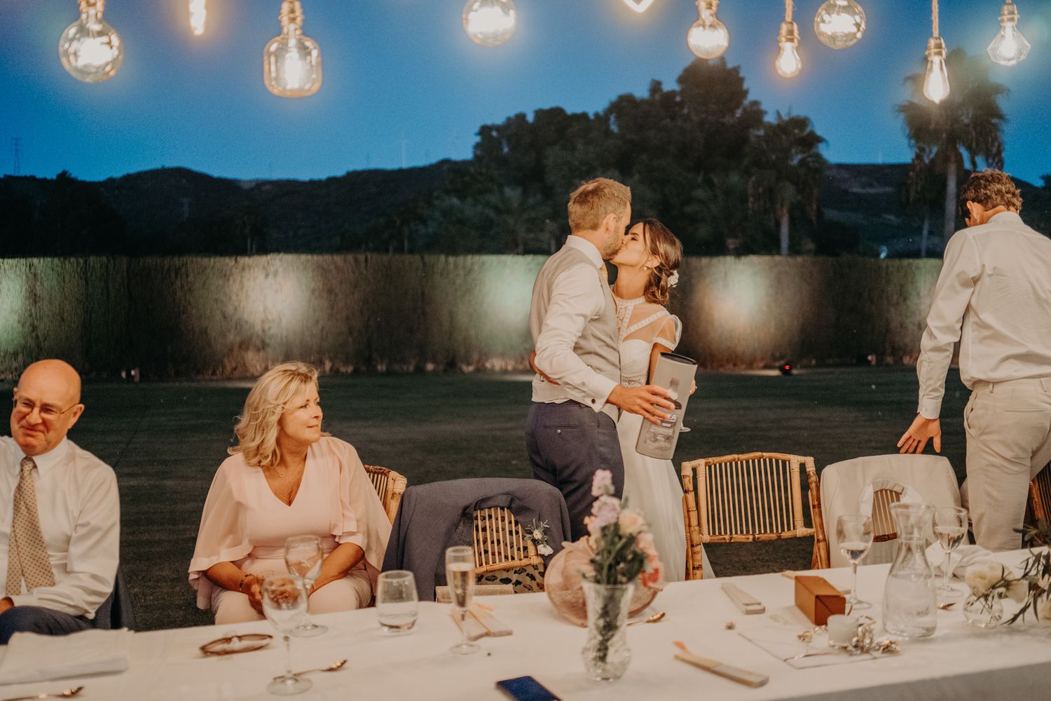 Evening outdoor reception with hanging bulb lights and dinner tables set against a mountain backdrop at dusk.