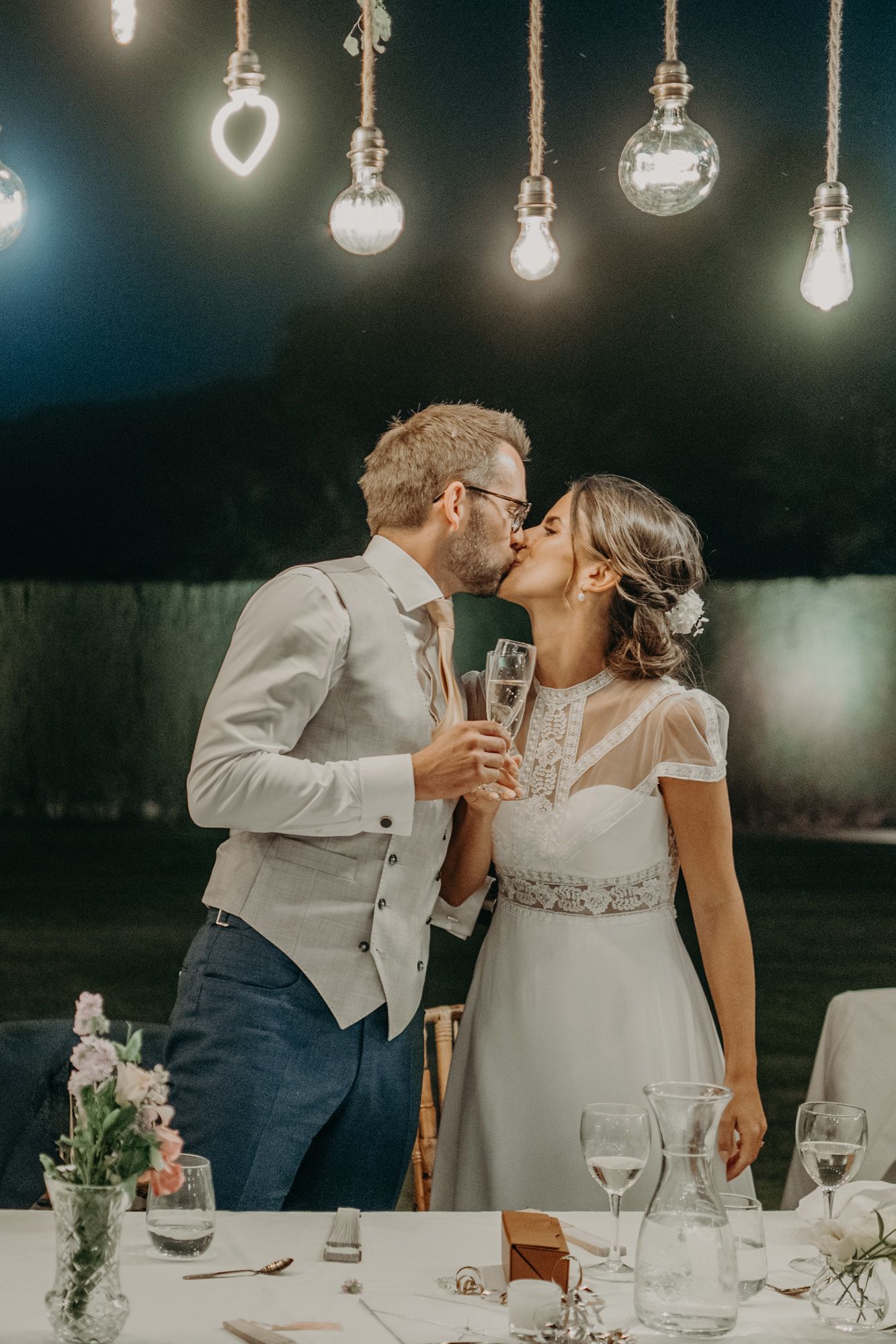 A romantic moment at a wedding reception with dangling Edison bulbs overhead and champagne glasses on the table.