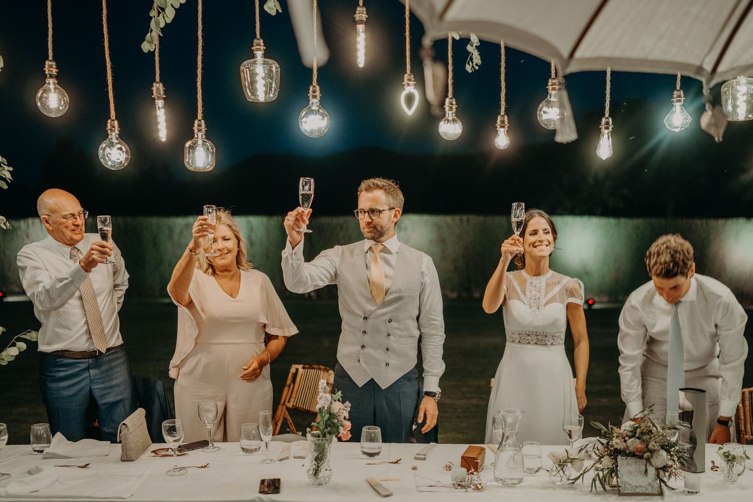 Wedding party stands at reception table holding up drinks for a toast under hanging vintage light bulbs.