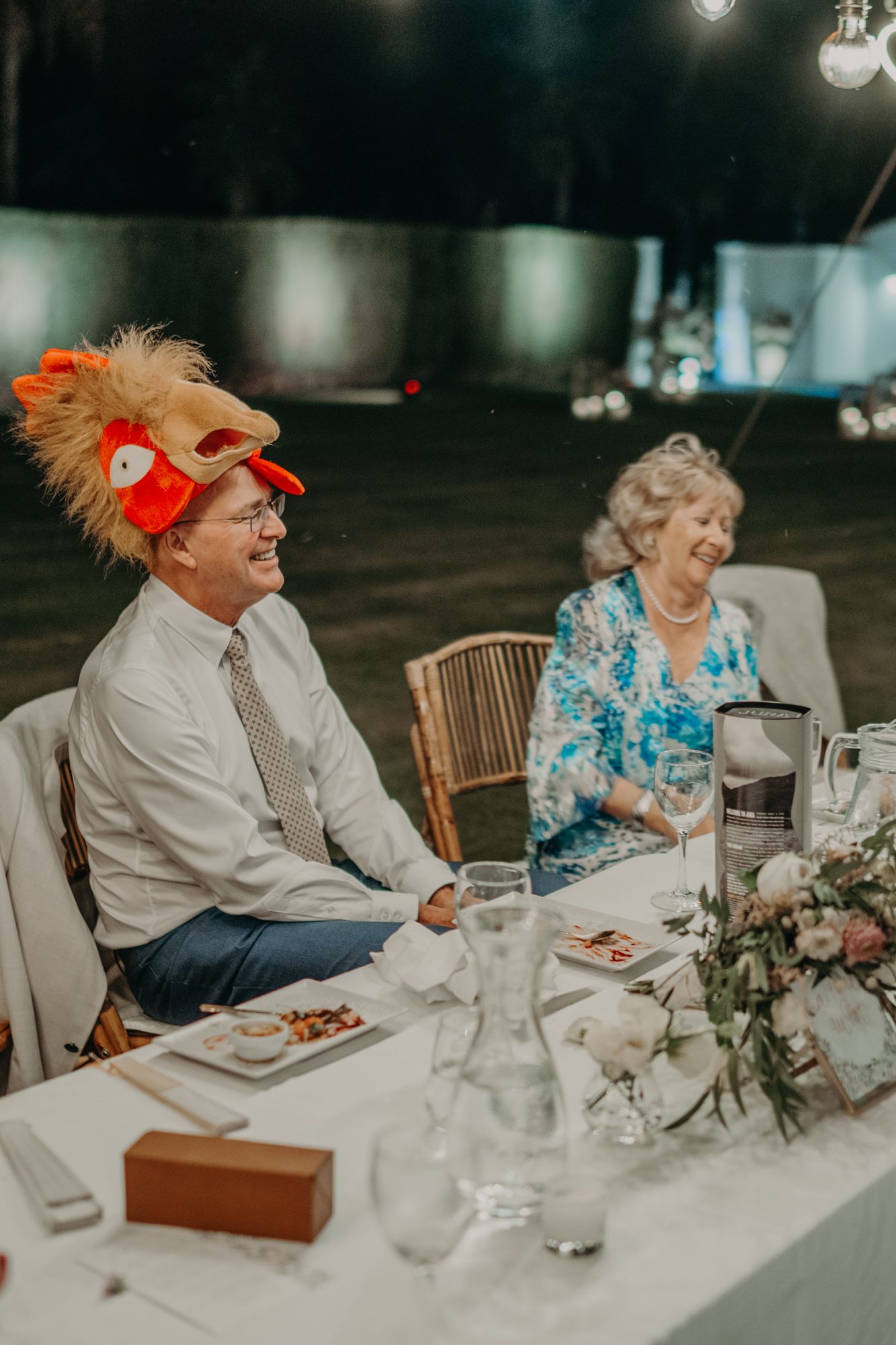 Wedding guest wearing a quirky red novelty hat sits at an elegantly decorated reception table.