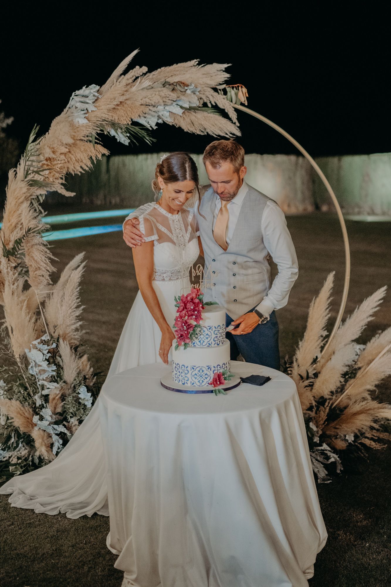 A couple cutting their wedding cake beneath an elegant arch decorated with pampas grass and floral arrangements.